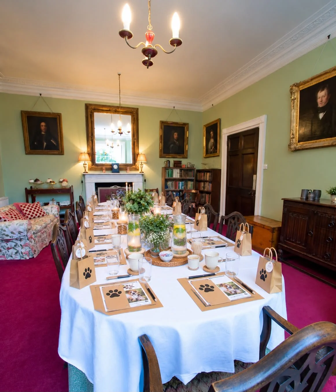A dining room set up for a party with a long table covered with a white tablecloth, decorated with greenery, glasses, plates, and small brown paper bags with paw print designs. There are water pitchers with lemon slices and mint, cups, and cutlery. The room has green walls, framed portraits, a mirror, a bookshelf, a fireplace, and vintage wooden furniture, with a chandelier hanging from the ceiling.
