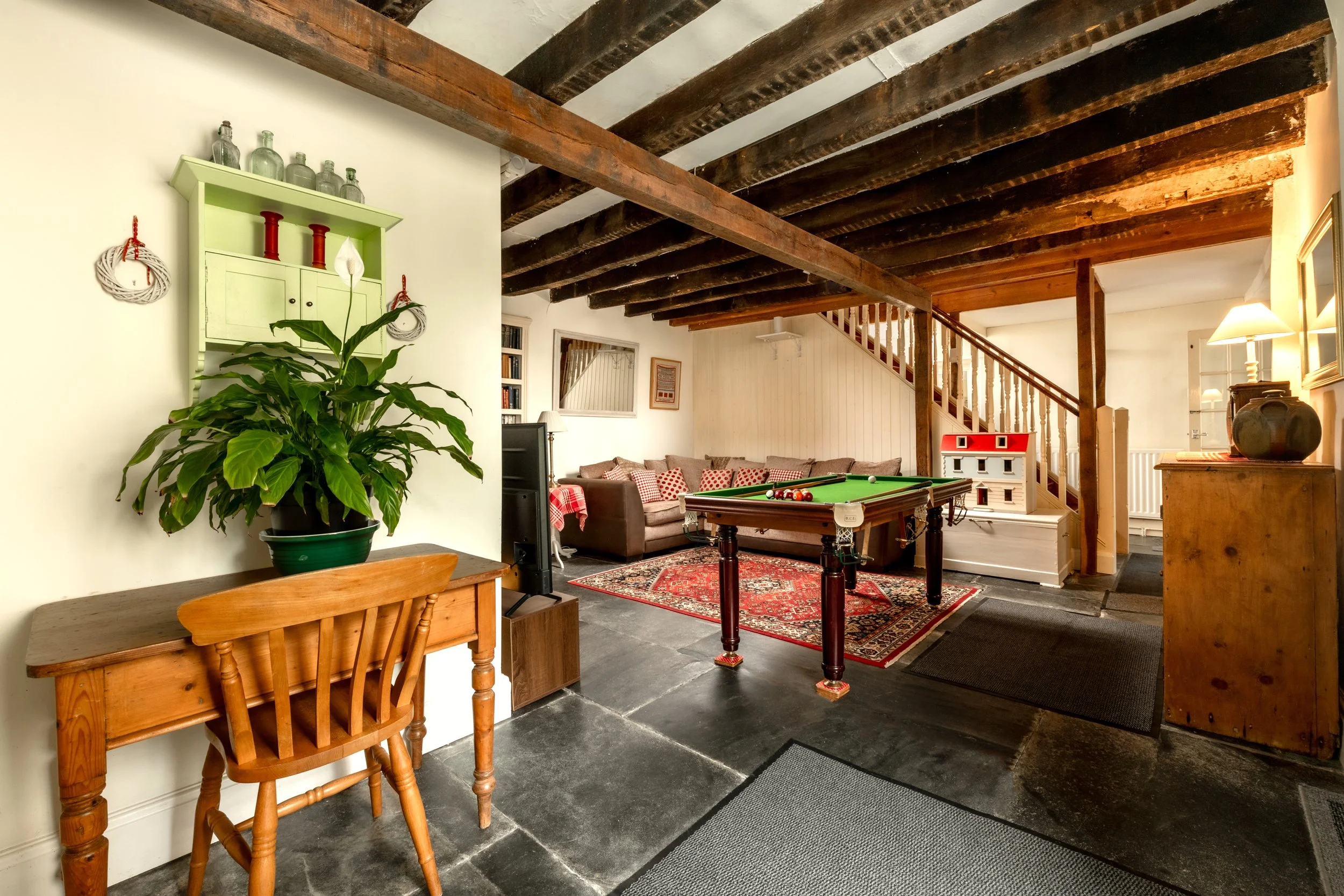 Living room with wooden beams on white ceiling, a large potted plant near a light green wall shelf with jars, a dining table, upholstered sofa with red-patterned cushions, pool table, red and white dollhouse, & warm lighting from lamps at Parnacott.