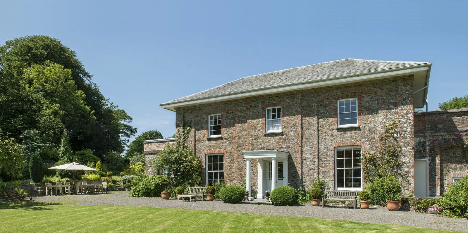 A large brick house with white windows and a small front porch, surrounded by a well-maintained lawn and garden with potted plants, trees, and outdoor seating on a clear day at Parnacott house.
