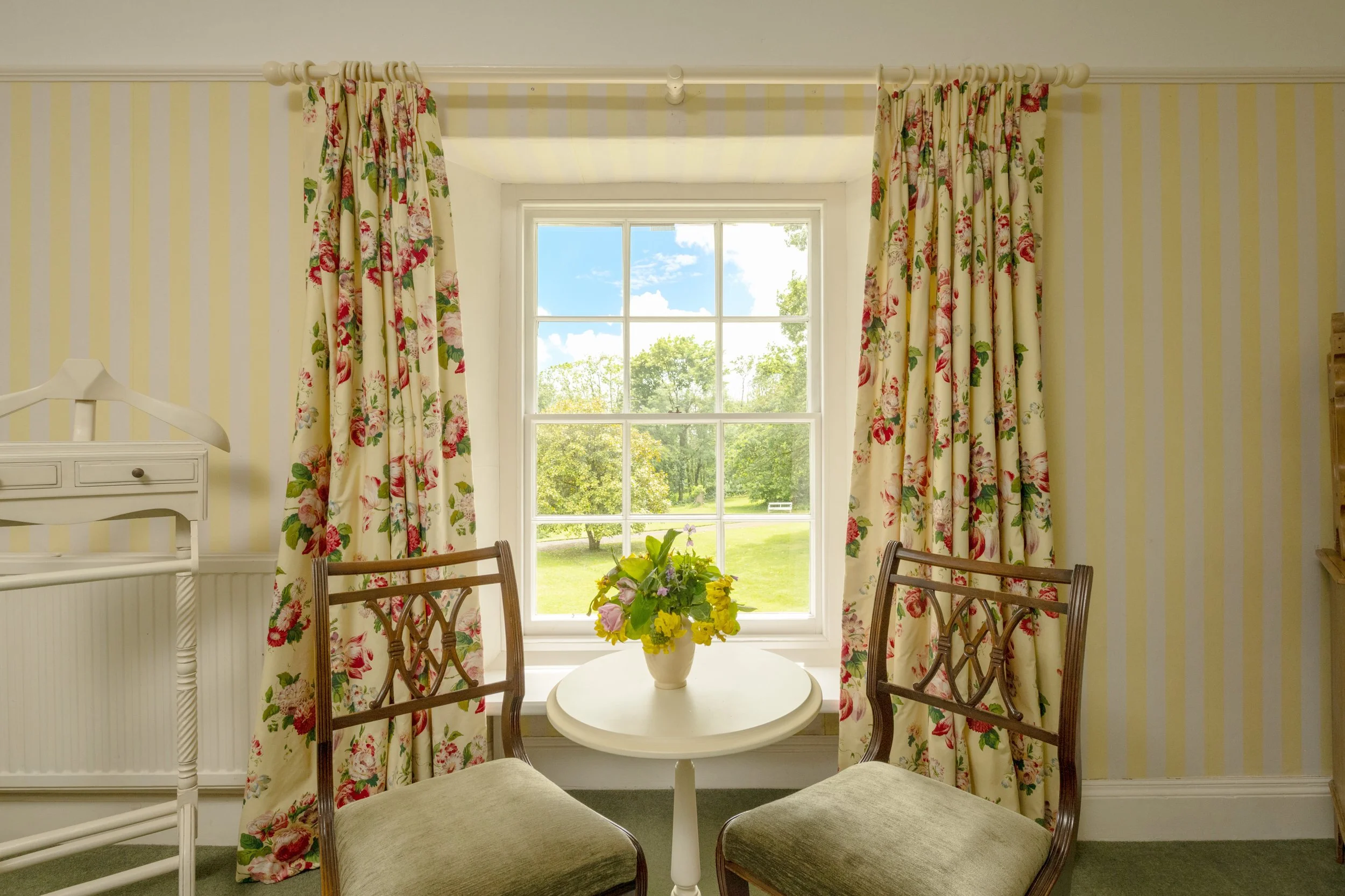 A bright room with yellow striped wallpaper, floral curtains, a white round table with a flower vase, and wooden chairs, overlooking a garden through a large window at Parnacott house. 