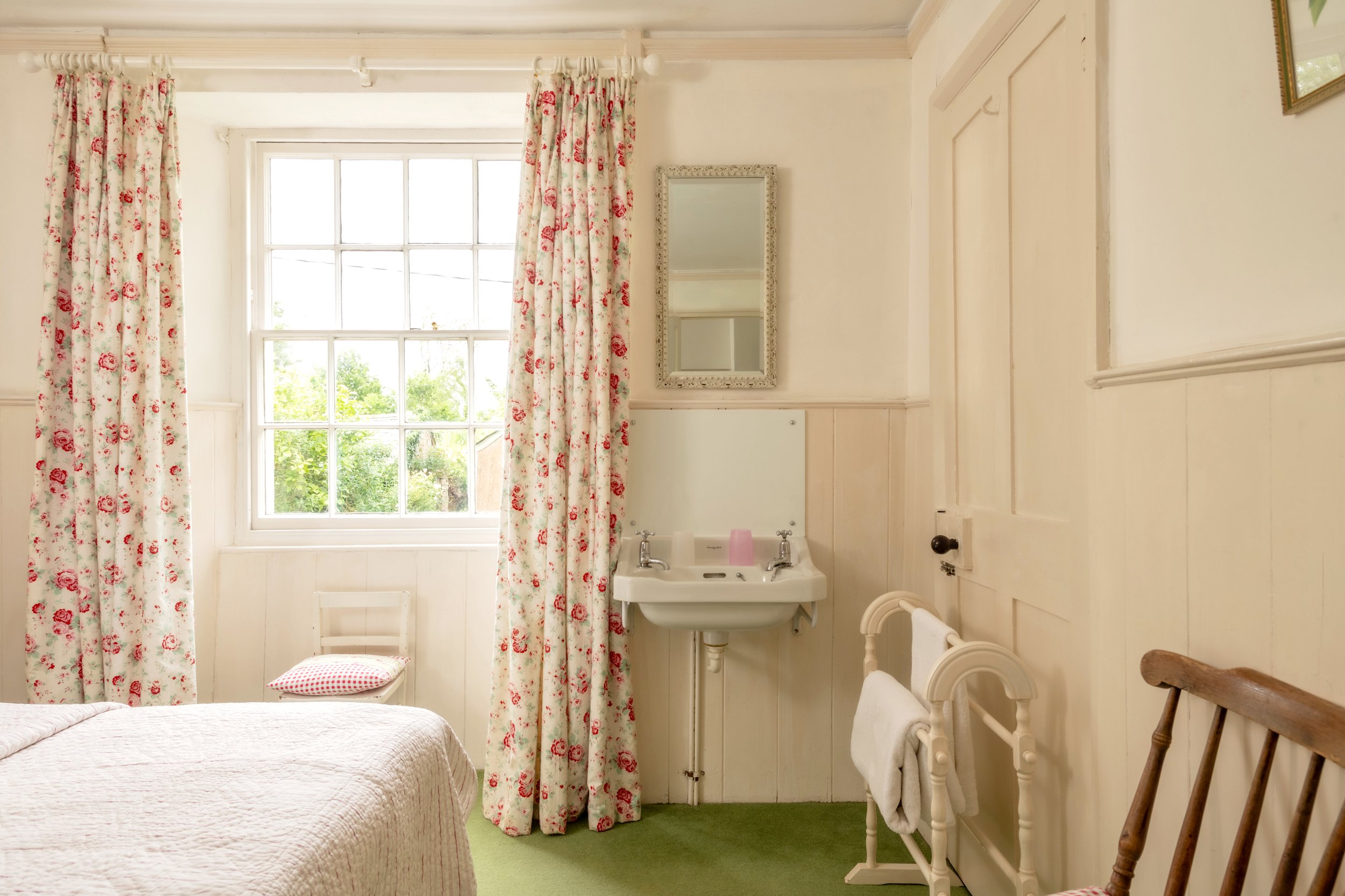 A cozy bedroom with a large window, floral curtains, a white sink with a mirror above, a wooden chair, and a bed with a quilted blanket at Parnacott house. 