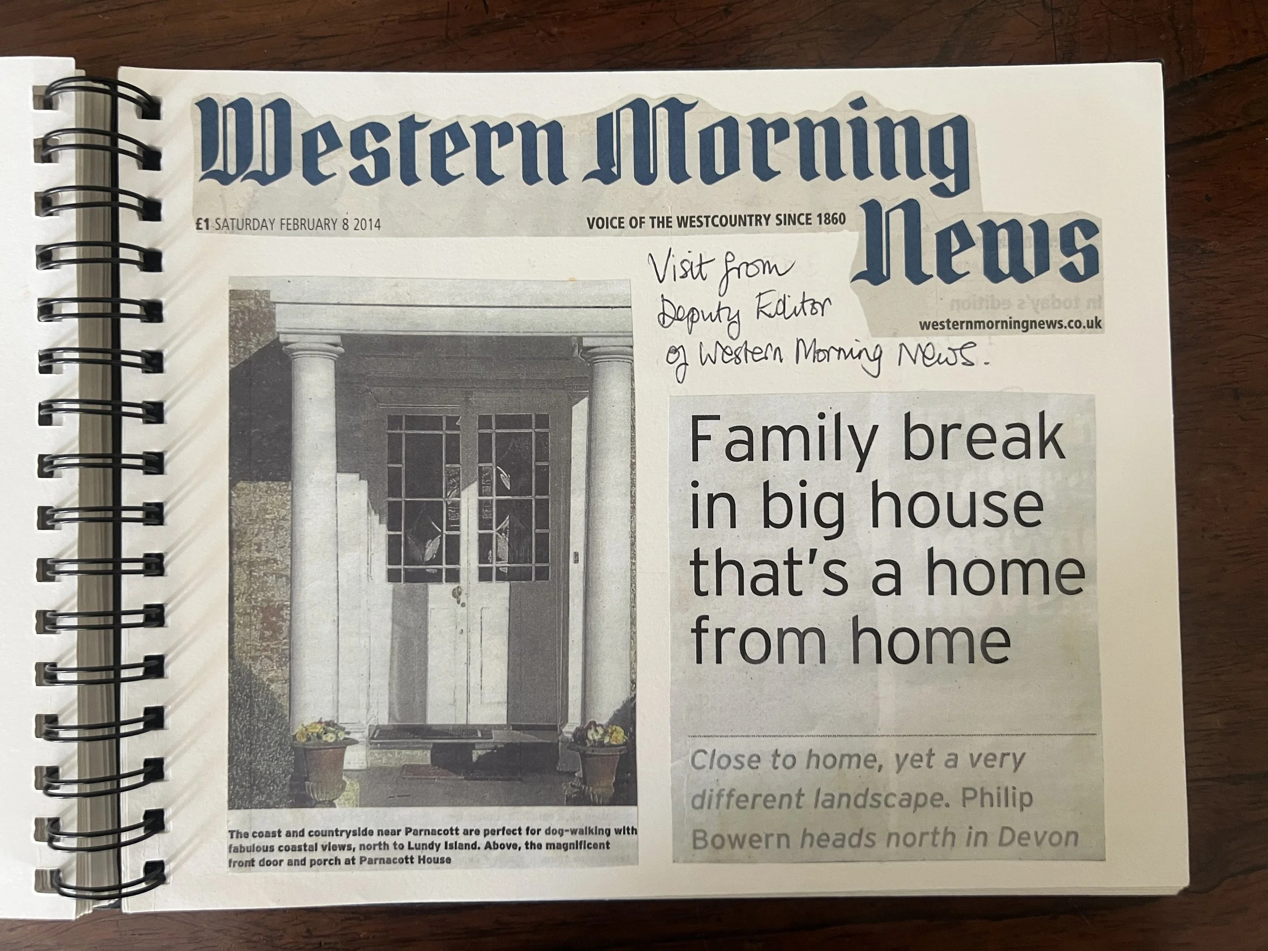 Page from a spiral-bound newspaper or magazine titled "Western Morning News" dated Saturday, February 8, 2014. The headline reads "Family break in big house," with a black and white photo of the front door and porch at Parnacott House, showing columns and a dog-walking pathway. Handwritten note says "Visit from Deputy Editor of Western Morning News."