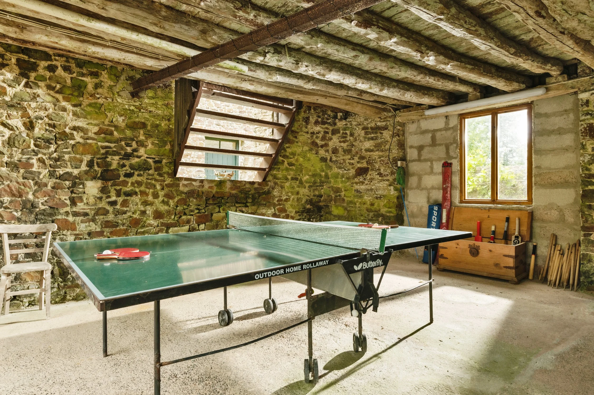 Indoor rustic room with exposed stone and brick walls, wooden ceiling beams, large window with natural light, a table tennis setup with paddles and balls, and badminton rackets stored in a wooden box at Parnacott house.