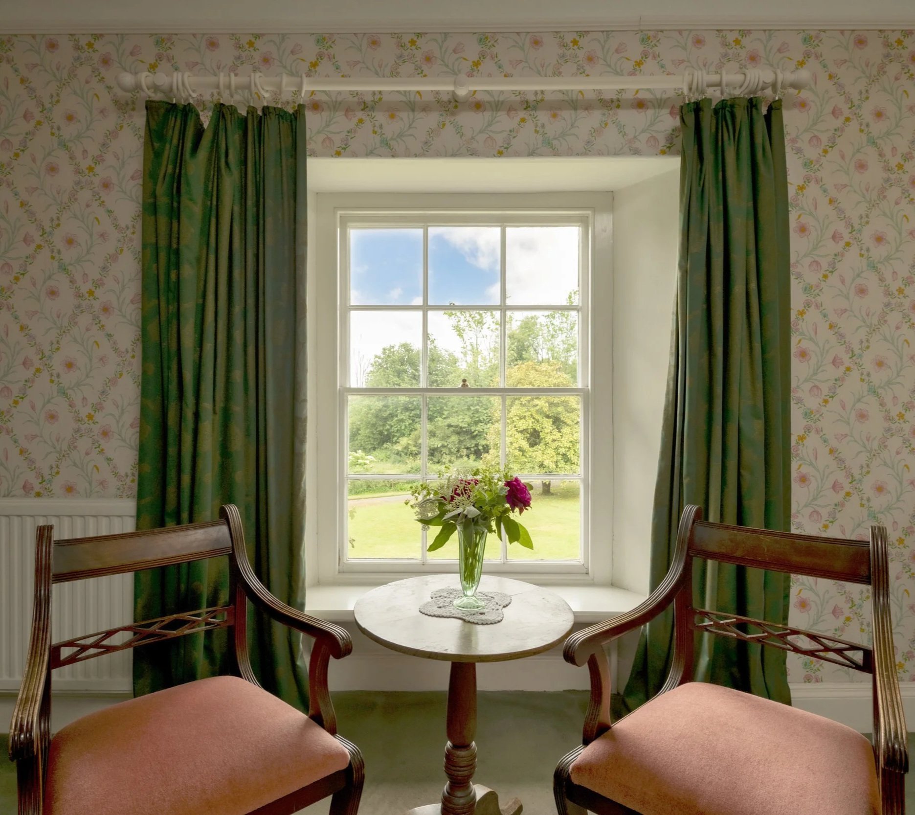 A cozy dining area with a round white marble table, a clear glass vase with pink and white flowers, and two pink-cushioned wooden chairs. Behind the table, there is a large window with white trim, revealing a green outdoor landscape with trees. Flanking the window are green curtains, and the wall behind features floral wallpaper with pink and yellow flowers and green vines