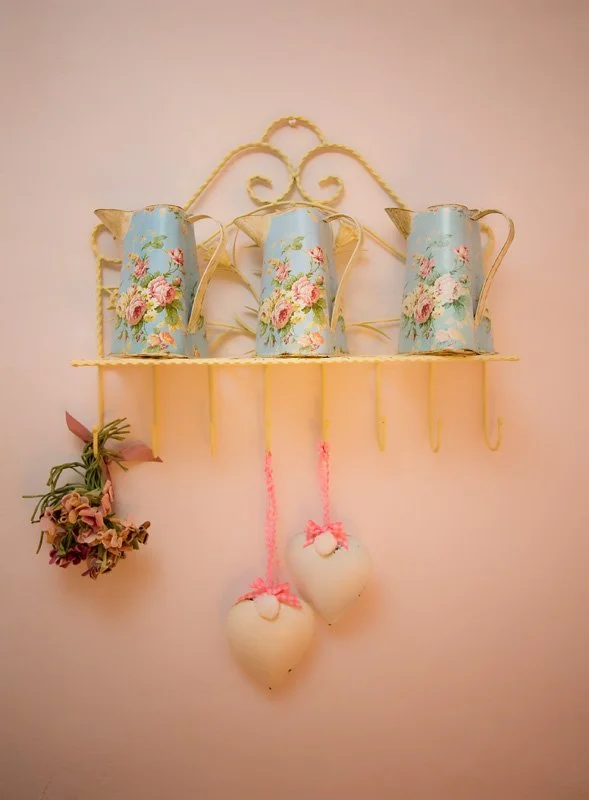 Decorative wall shelf with three vintage-style blue watering cans with floral patterns, hanging pink heart-shaped ornaments tied with pink yarn, and a bouquet of flowers hanging from the left side at Parnacott house. 