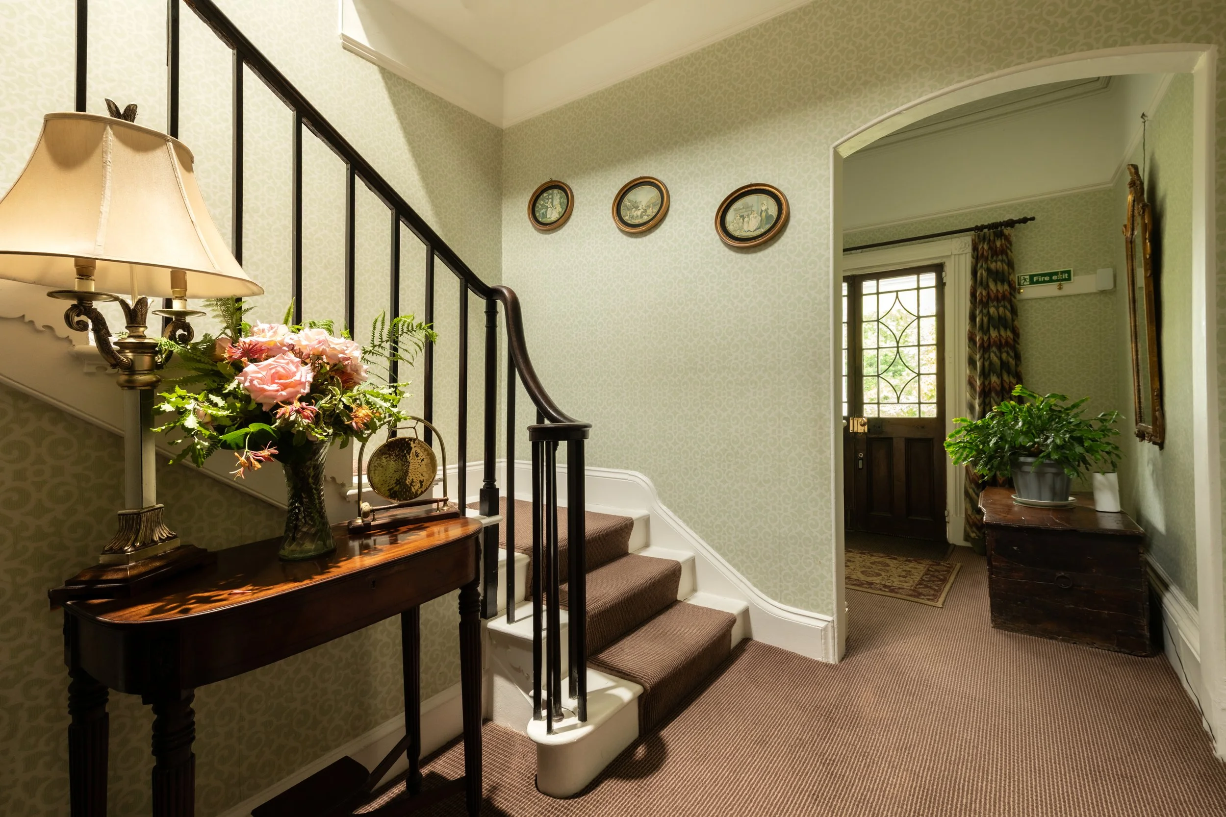 Interior view of a house entryway with stairs, a lamp, floral arrangement, picture frames on the wall, a green patterned wall, and a door with window panes and a curtain at Parnacott house.