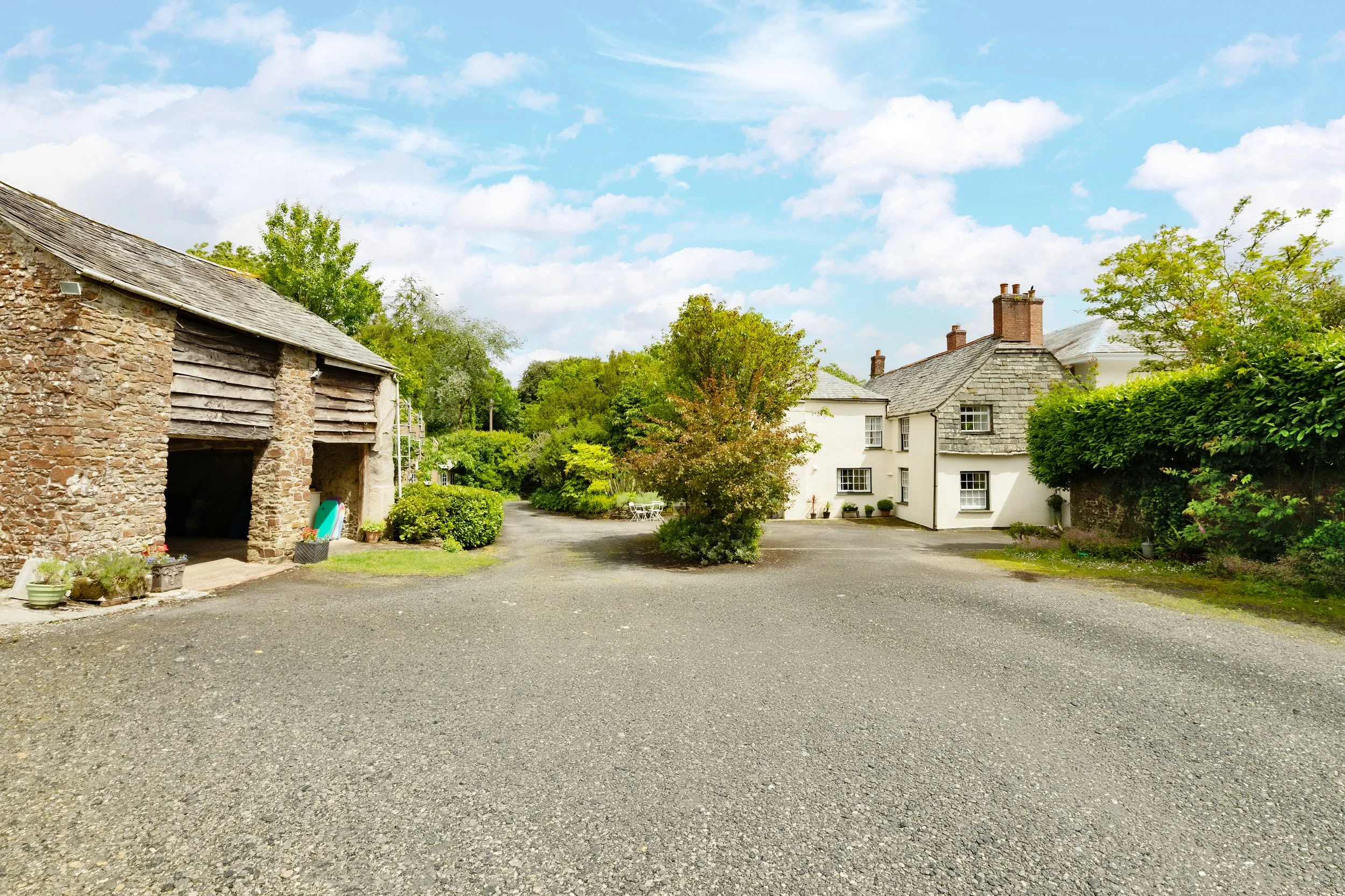 A rural scene with a gravel driveway leading to classic houses with stone and white exteriors, surrounded by lush green trees and bushes, under a partly cloudy sky at Parnacott house.