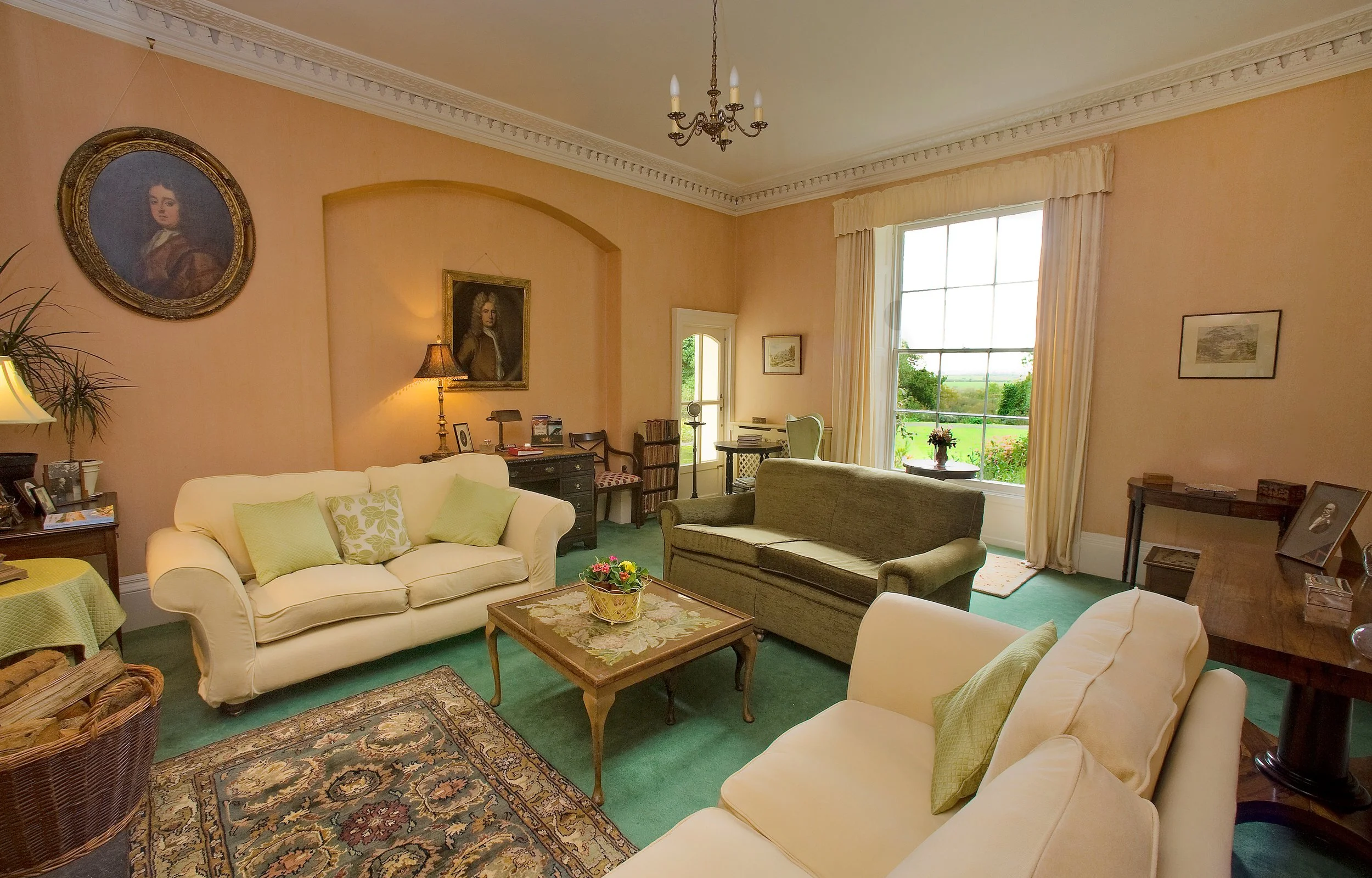 Living room with peach walls, large window with cream curtains, chandelier, portrait paintings, cream and green sofas, wooden coffee table with a flower arrangement, various side tables, lamps, and framed photographs at Parnacott house.
