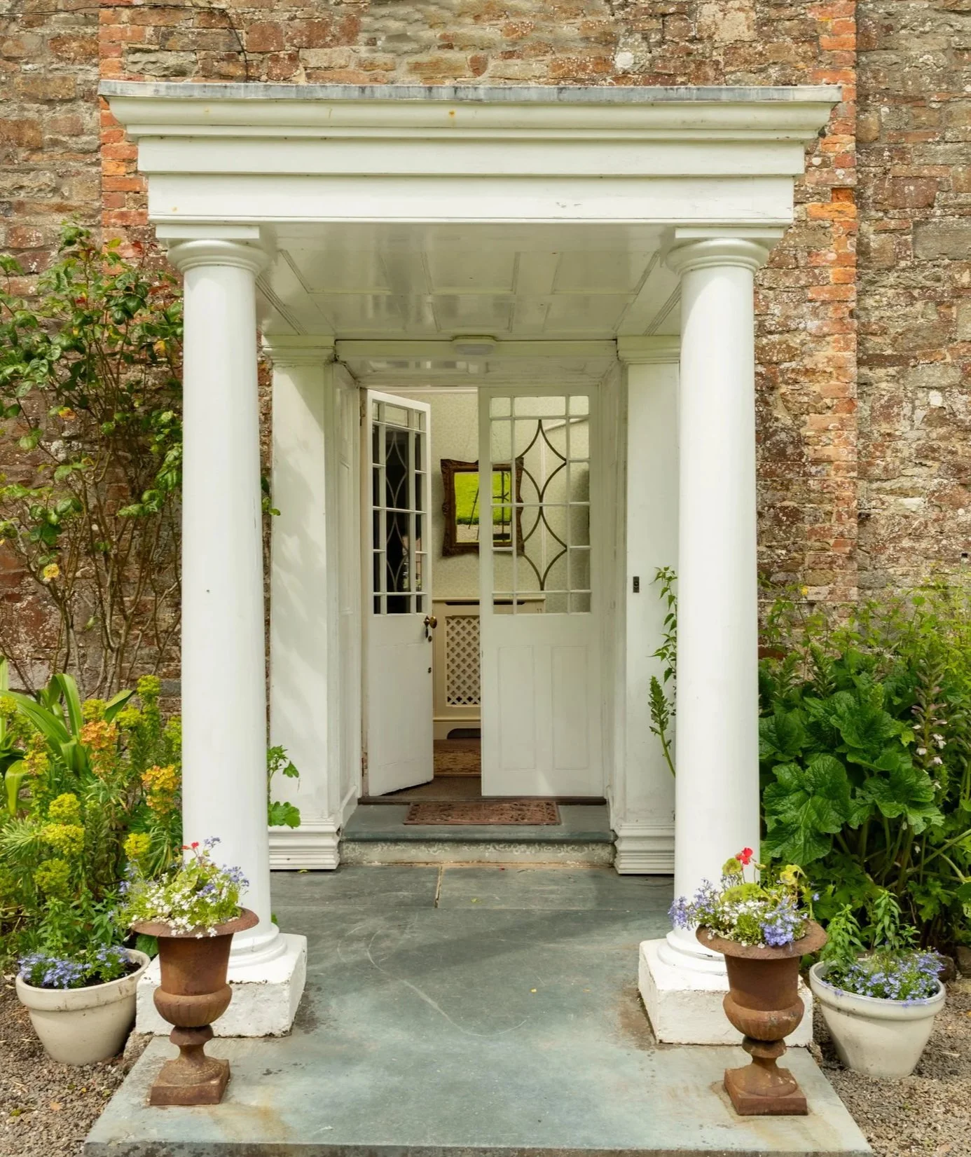 White porch with columns, open glass-paneled door, potted flowers, and a brick wall background at Parnacott house.