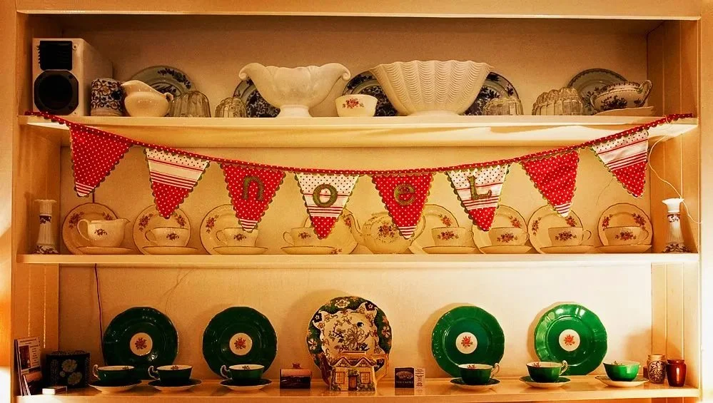 Kitchen display shelf decorated for Christmas with vintage dishes, plates, bowls, cups, and a festive banner spelling out "NOEL" with red and white pennants at Parnacott house.