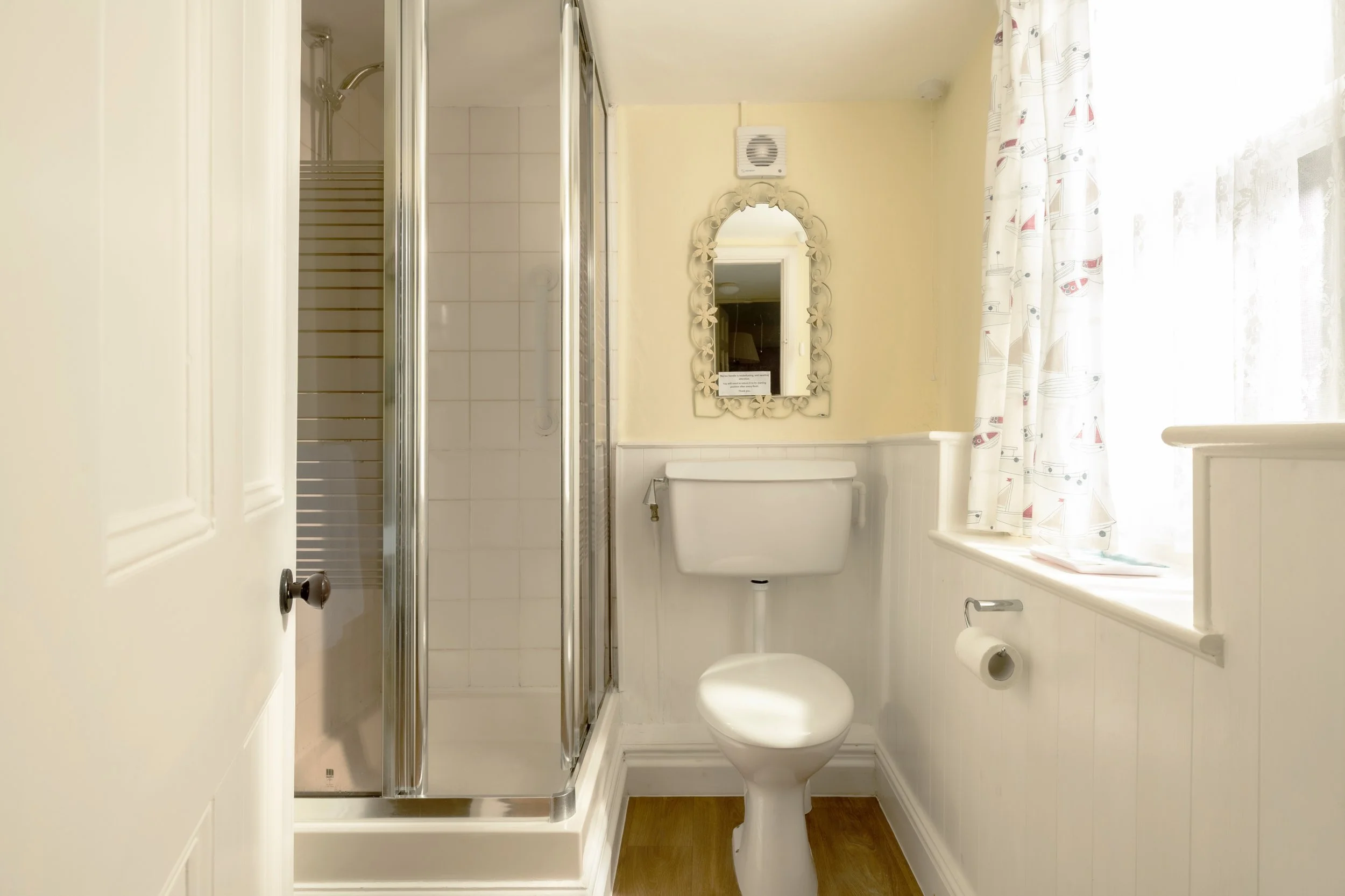 Small bathroom featuring a toilet, a window with patterned curtains, a mirror with floral frame, a shower enclosure, and a wall vent at Parnacott house. 