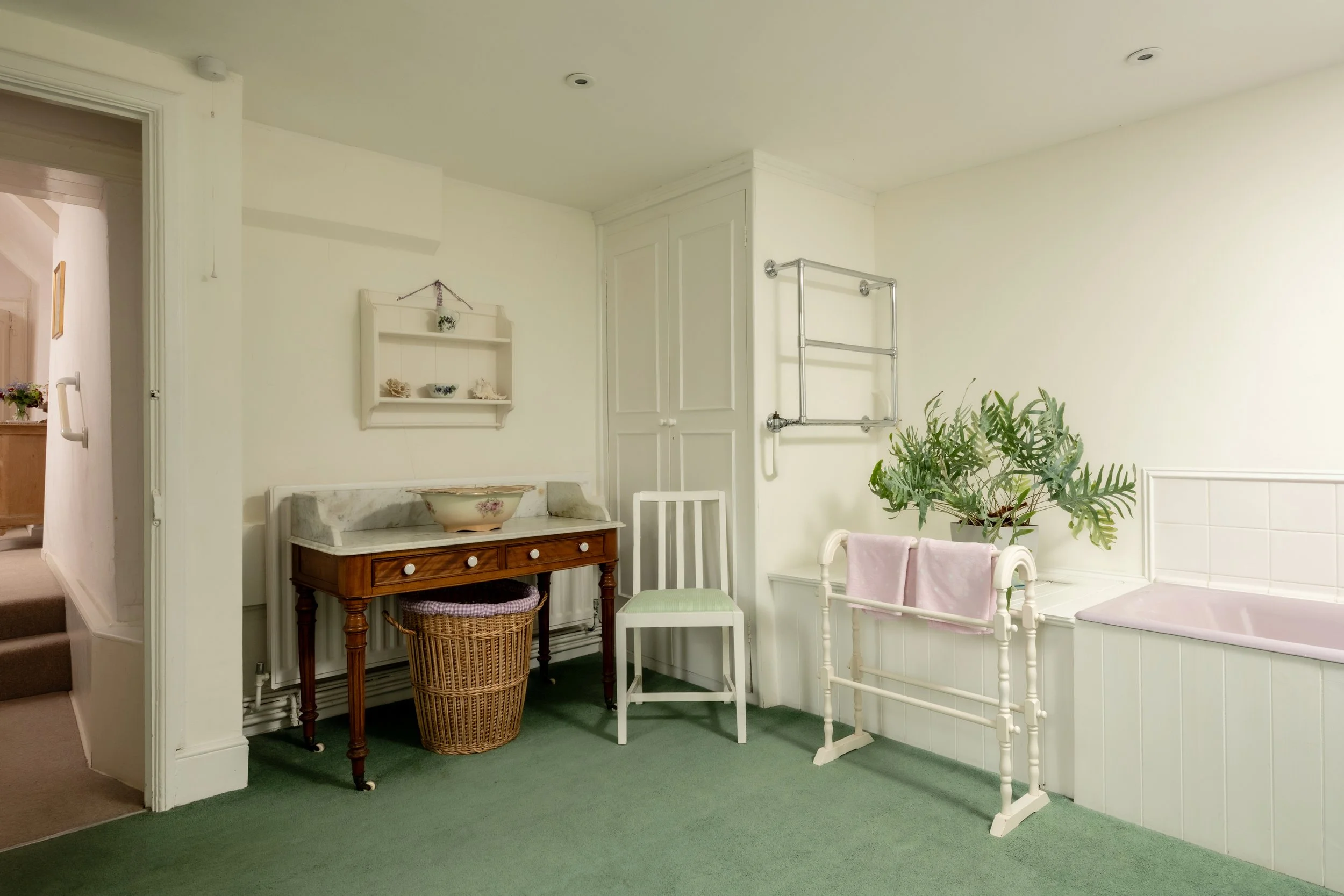 A cozy vintage-style corner in a home with white walls and green carpeting. It features a wooden table with curved legs, a wicker basket underneath, two upholstered chairs with pink covers, a white chair, a large potted fern, a white wall-mounted she