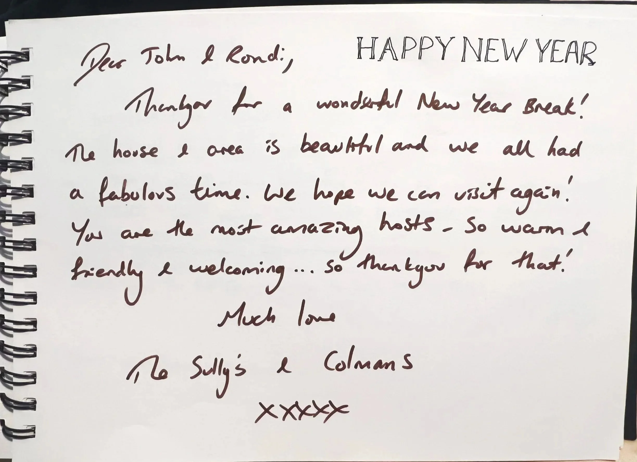 A handwritten note in a spiral-bound notebook with a white background. The message is addressed to John and Rondi and wishes them a Happy New Year. It thanks them for a wonderful New Year break, describes the house and area as beautiful, and expresses hope to visit again. The note ends with warm wishes and is signed by someone named Sally and Colman.