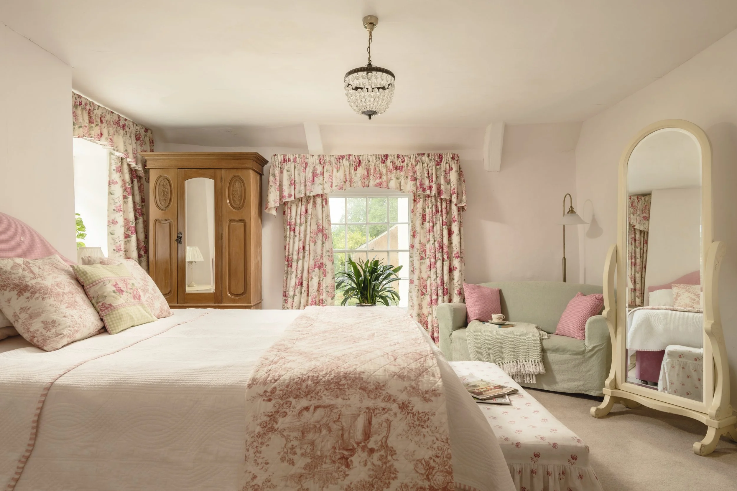 A cozy bedroom with two beds, floral curtains, an arched mirror, a wooden wardrobe, a chandelier, and a green armchair with pink pillows at Parnacott house. 