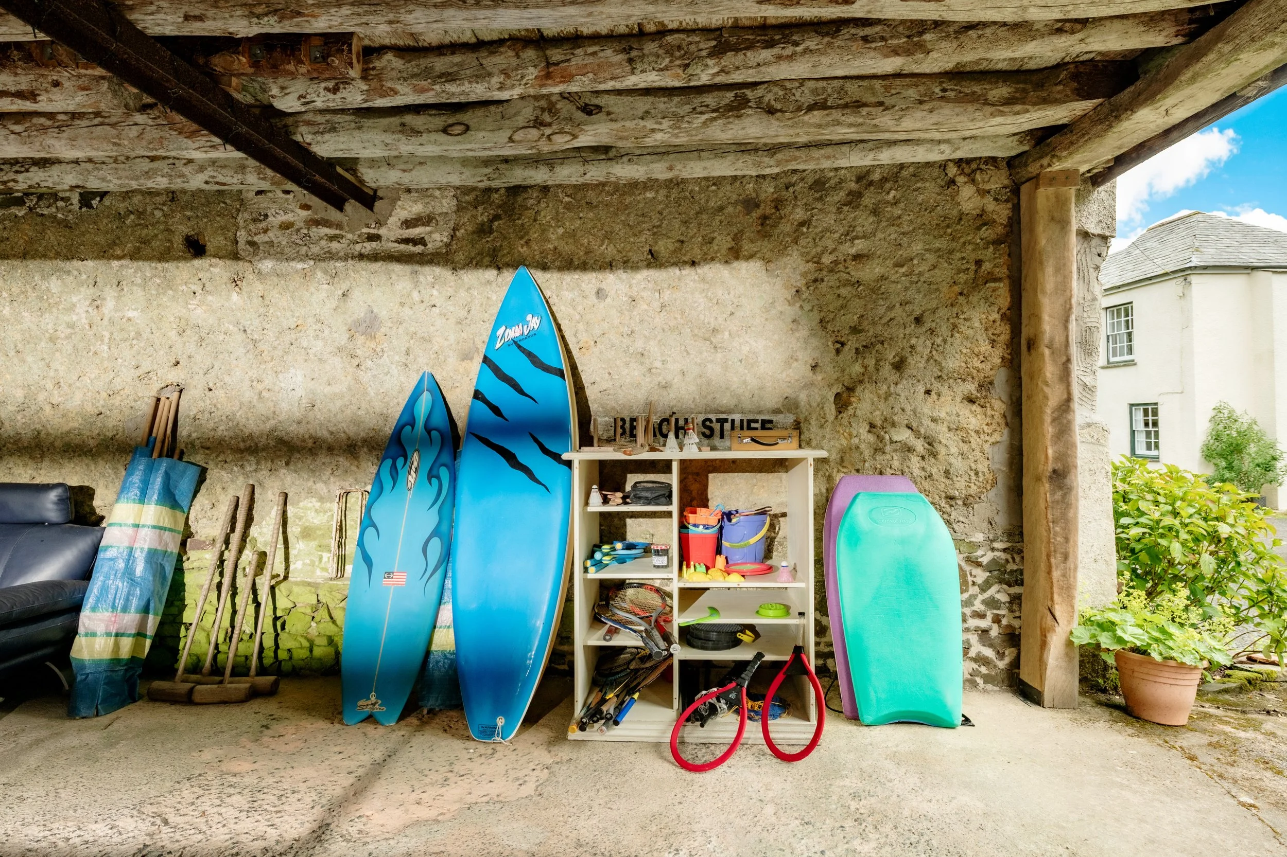 Garage with surfboards, paddles, and various outdoor toys and equipment stored on shelves and the ground, with a stone wall and a small window outdoor view at Parnacott house.