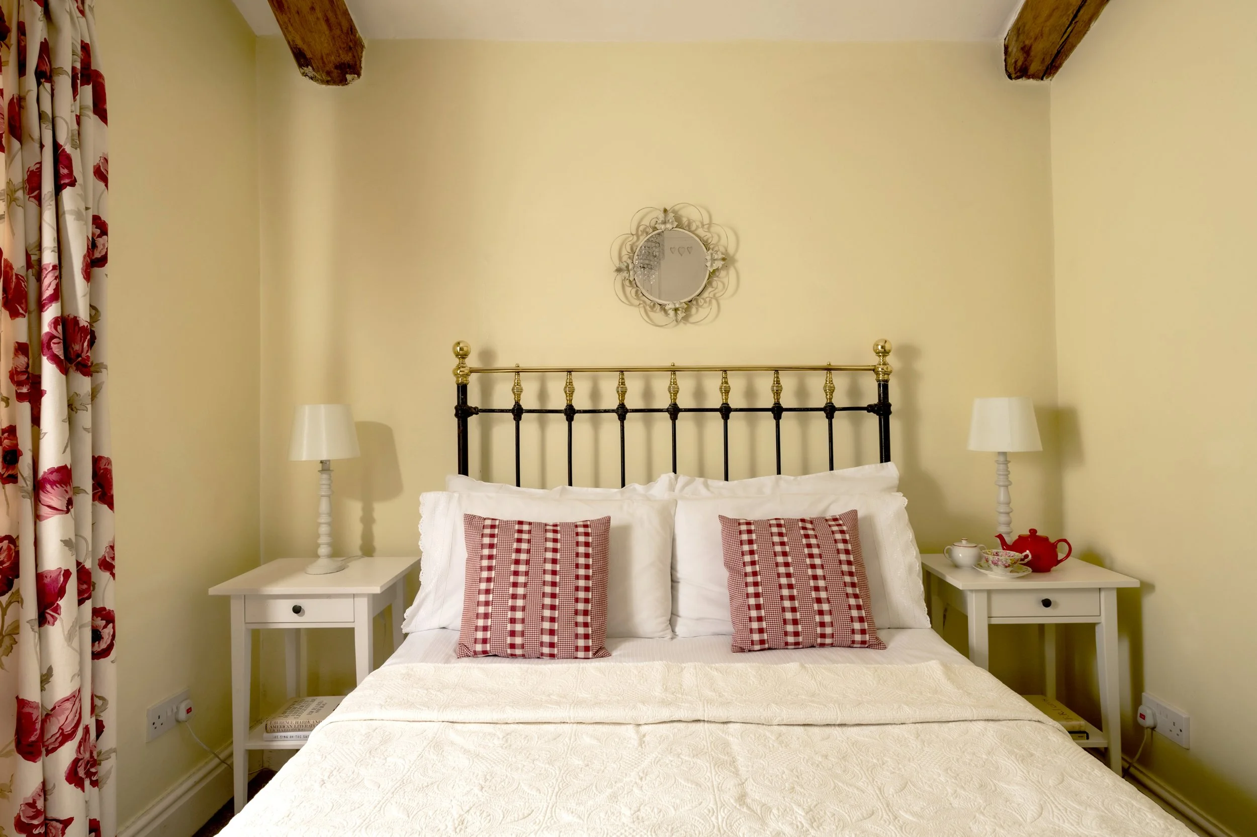 A cozy bedroom with a bed, red and white checkered pillows, white nightstands on each side with lamps, and a mirror on the yellow wall above the bed. Floral curtains are on the left side of the image at Parnacott house. 