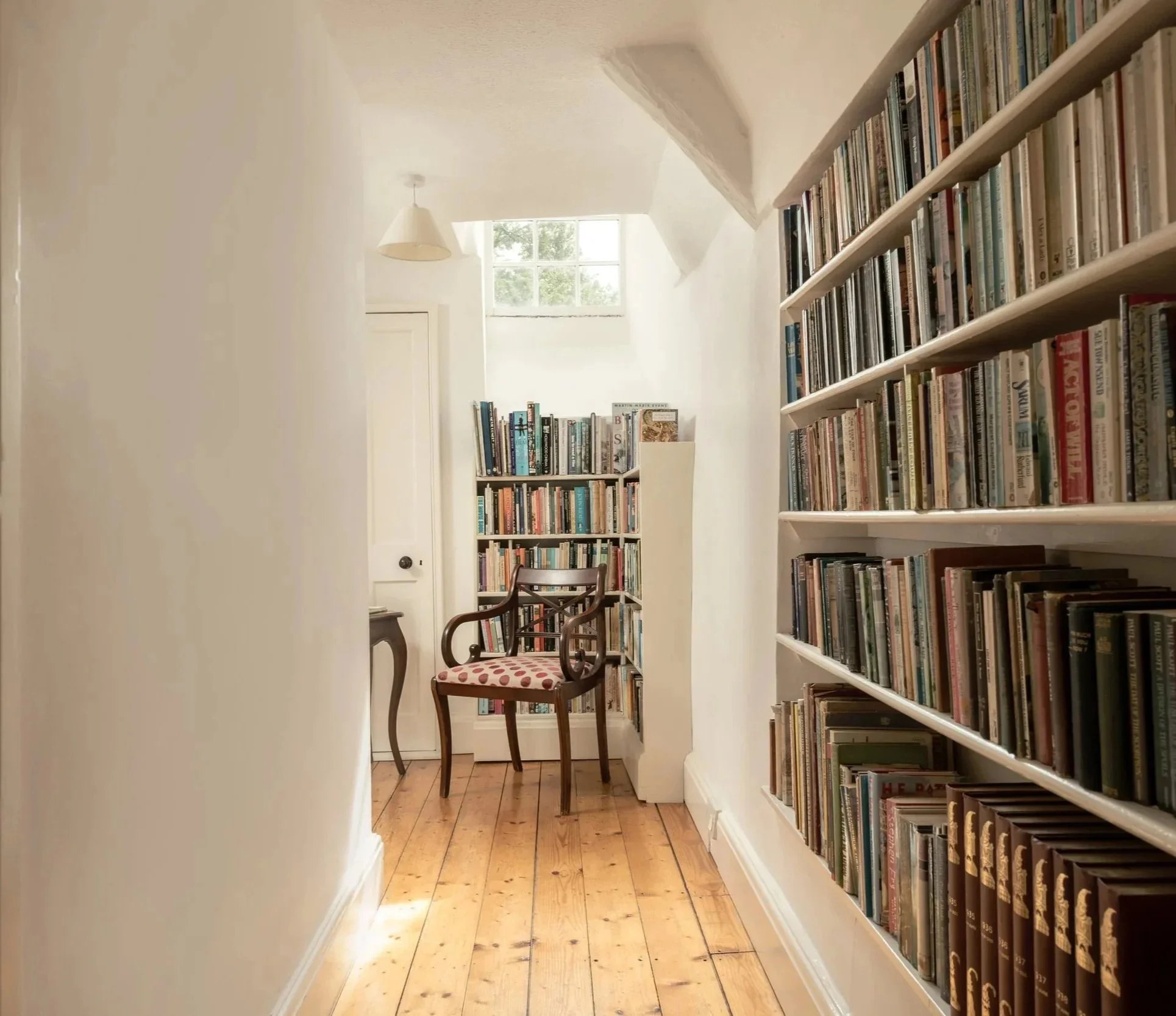 A cozy reading nook with white walls, a window letting in natural light, wooden flooring, and tall bookshelves filled with books. Two wooden chairs with cushioned seats are placed near the bookshelves at Parnacott house.