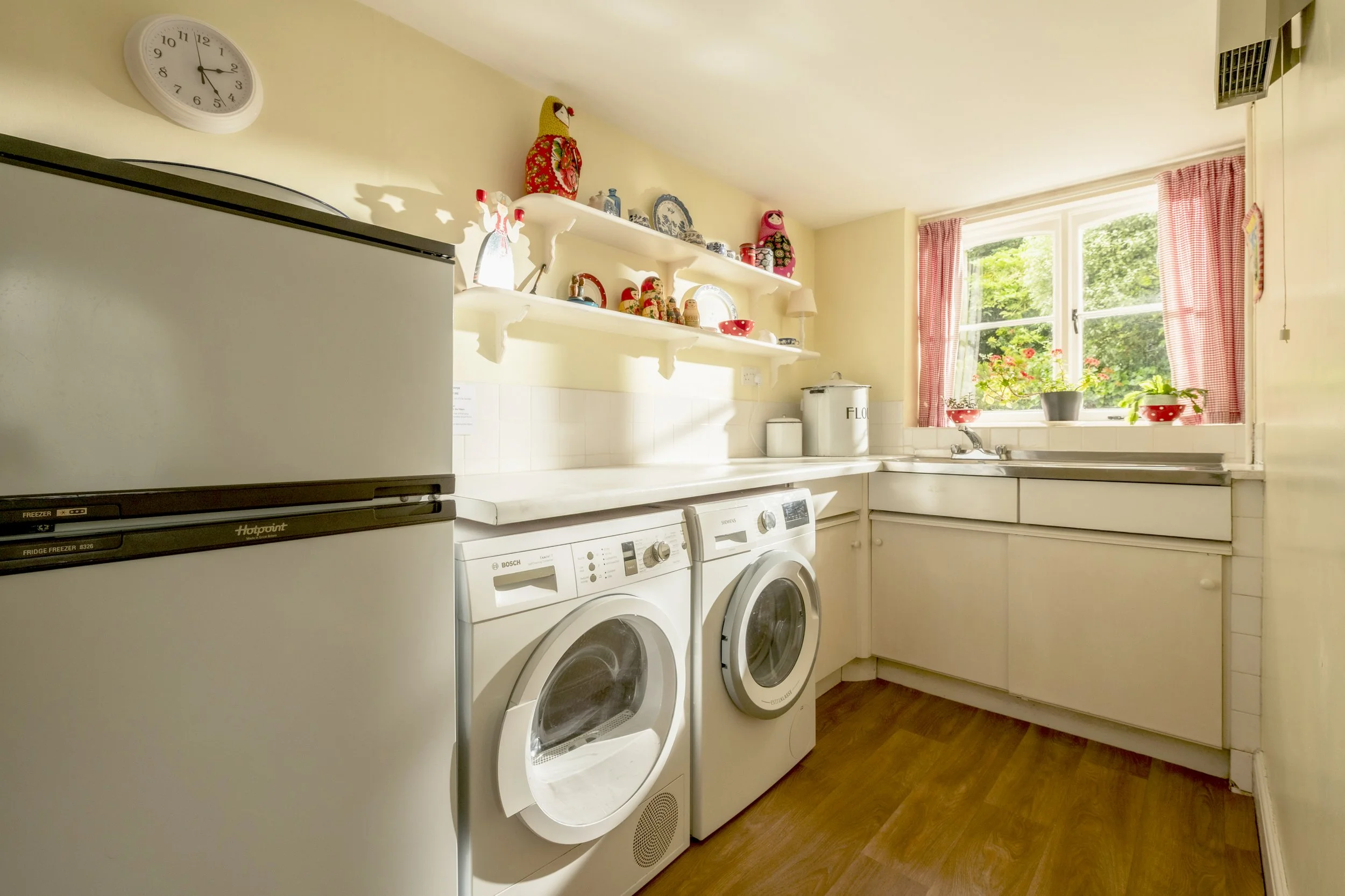 A laundry room with a white refrigerator, a washer, and a dryer beneath open shelves decorated with decorative dolls, plates, and jars. There is a window with red checkered curtains and several potted plants on the windowsill.