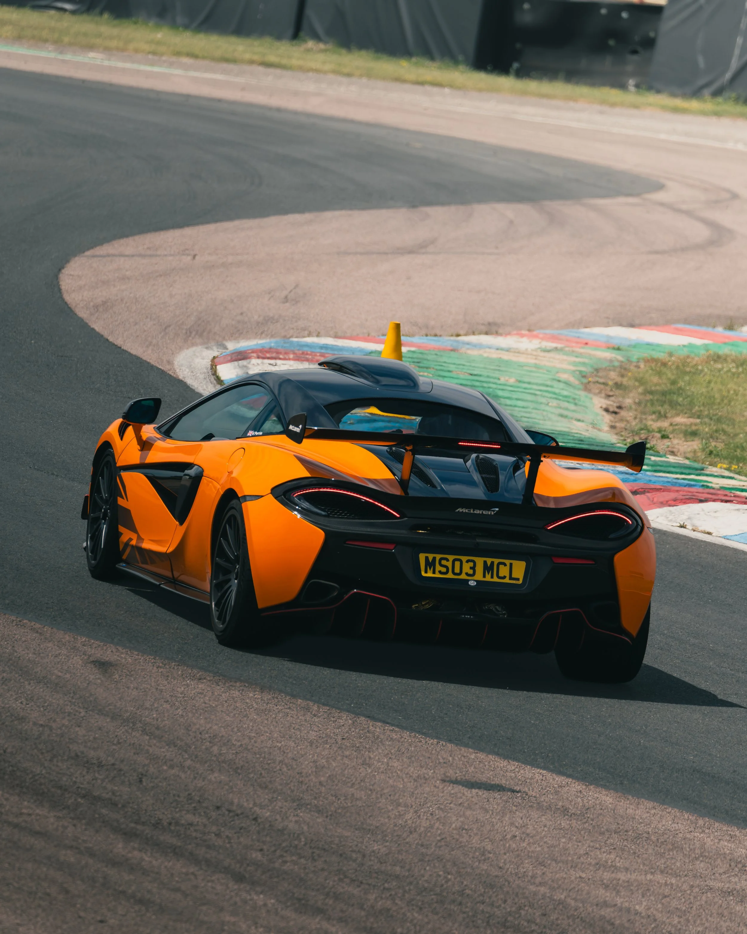 An orange and black sports car on a race track with a yellow caution cone on its roof. The race track has green, white, and red painted curbing and a curved asphalt section with tire marks. The background shows a grassy area and a black barrier.