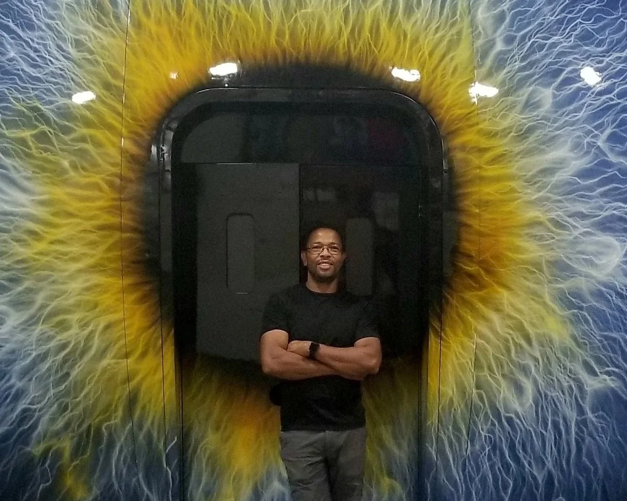 Man standing with arms crossed in front of a wall with a large eye mural, featuring a yellow and blue iris.