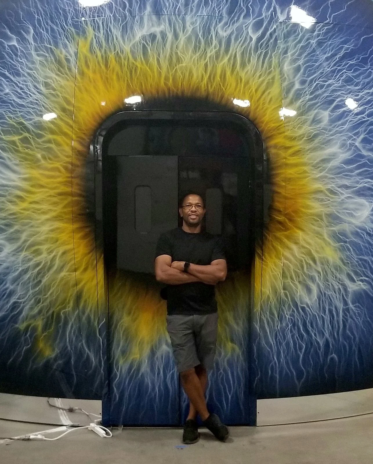 A man standing with arms crossed in front of a mural of a yellow eye, inside an elevator with black doors.