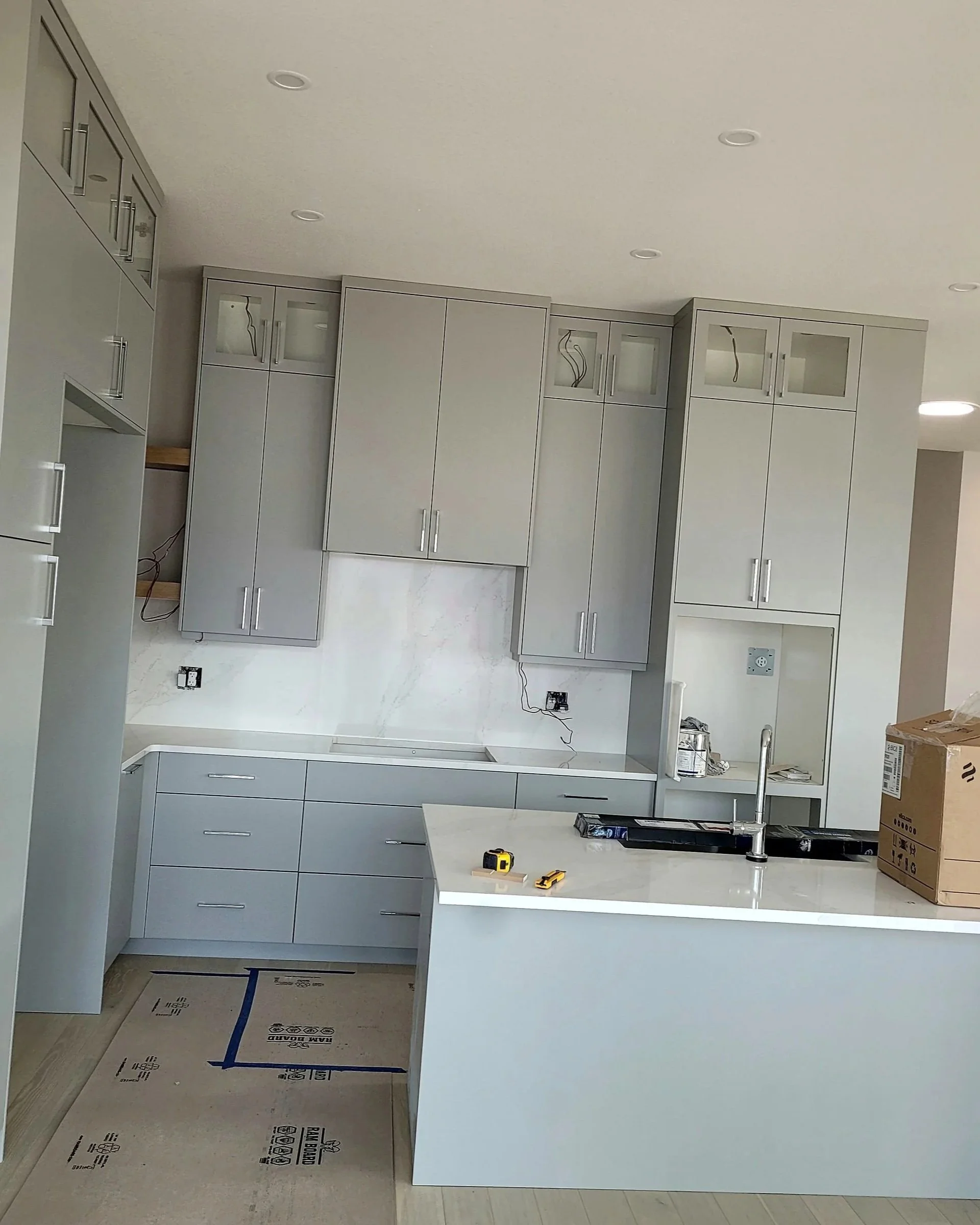 A kitchen in the process of being renovated with light gray cabinets, an island with a white countertop, a stainless steel faucet, and tools on the island. The floor is covered with protective paper.