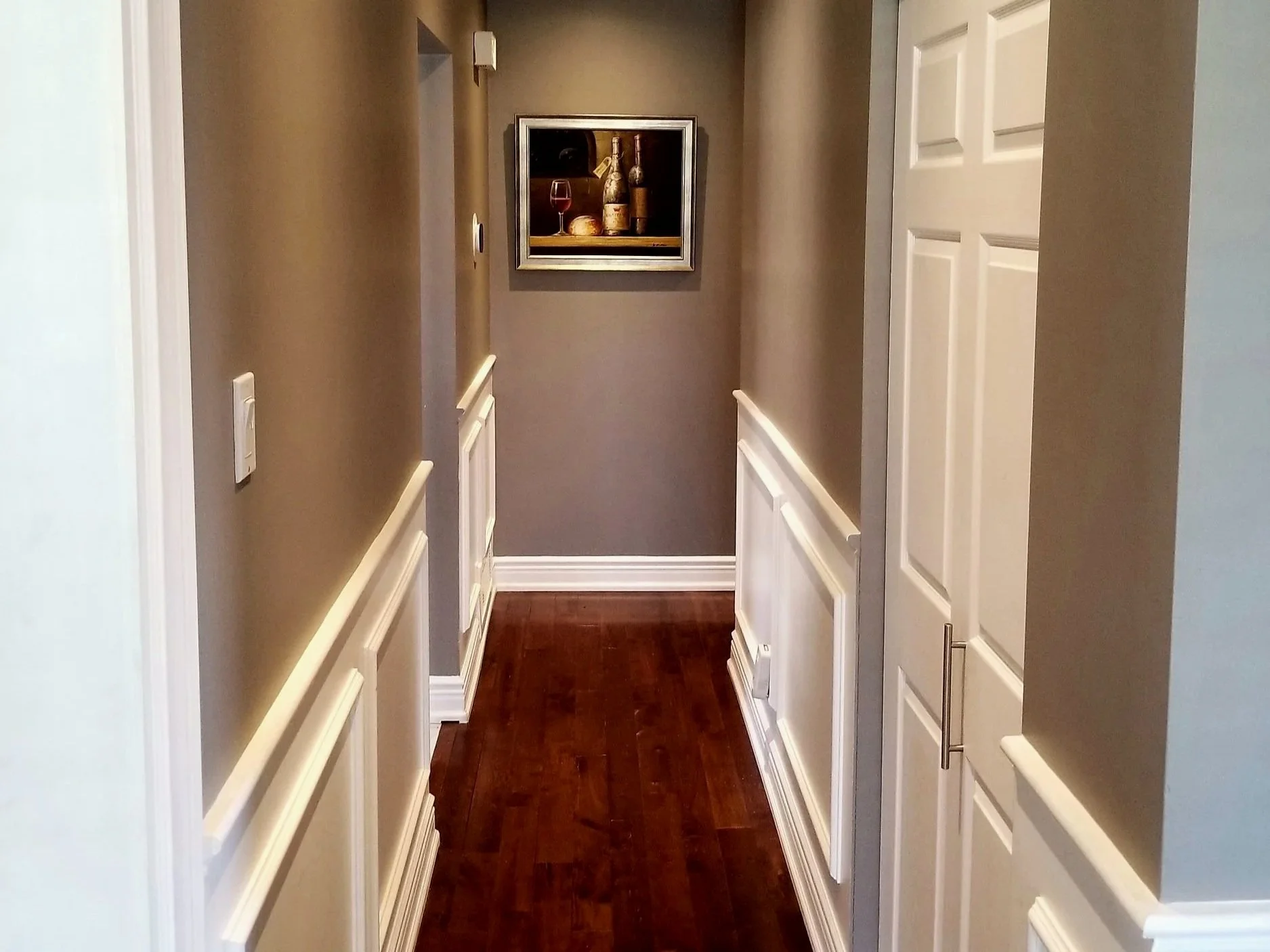 A narrow hallway with beige walls, white wainscoting, and a dark hardwood floor. At the end of the hallway, there is a framed still-life painting depicting bottles, a glass of wine, and cheese.