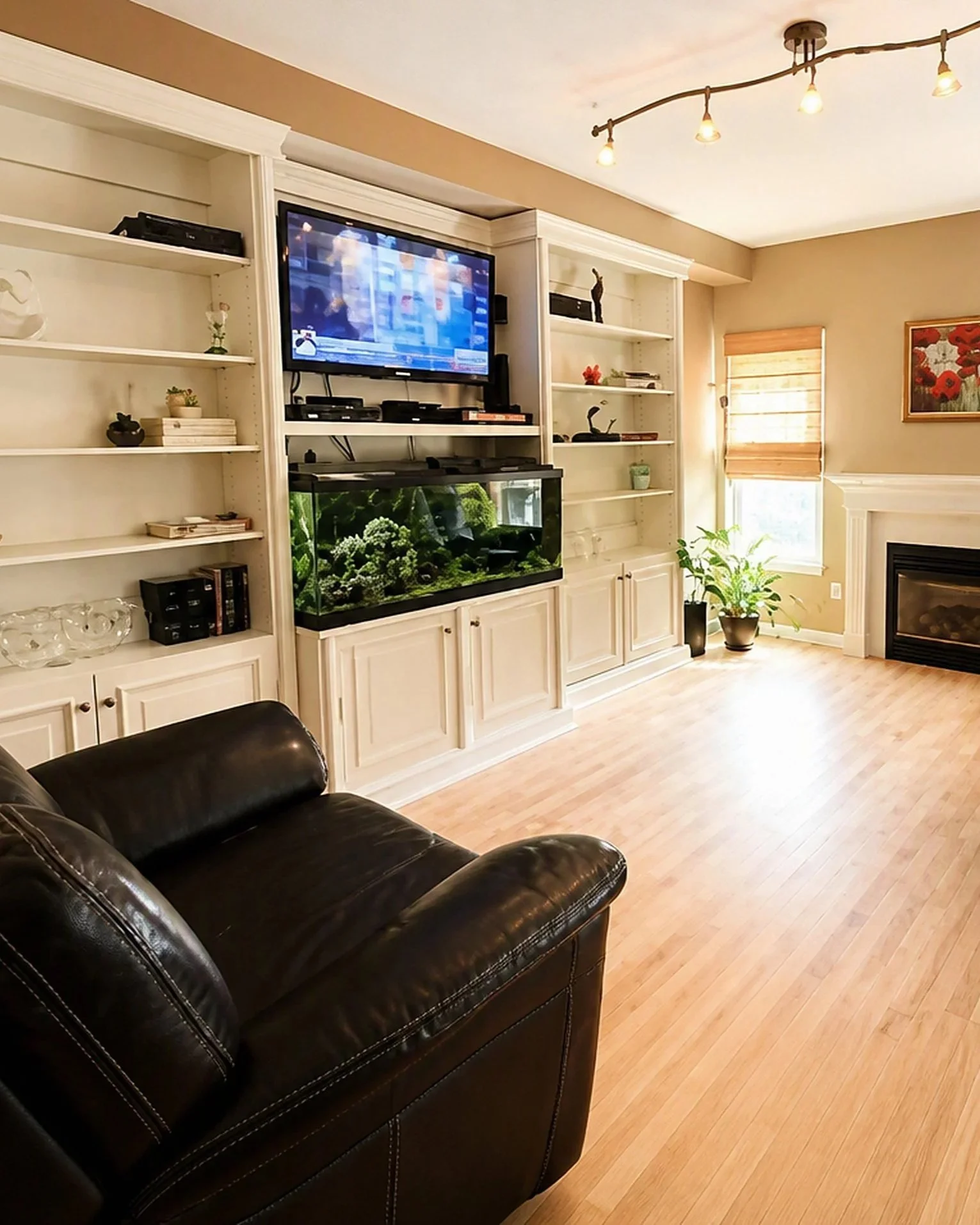 Living room with a black leather sofa, white built-in shelves, a large fish tank with plants, a wall-mounted TV, a window with blinds, room plants, and artwork on the wall.