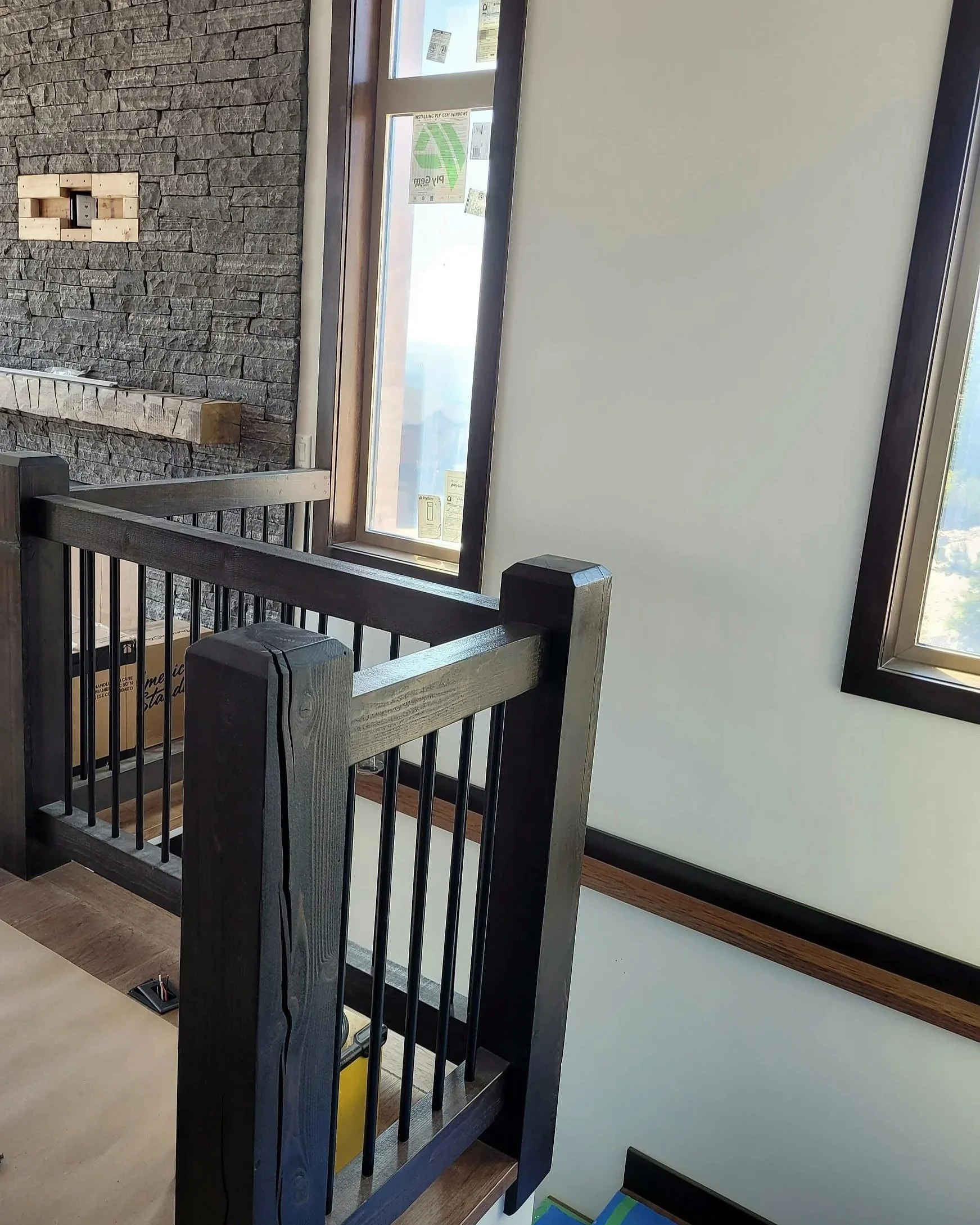 Interior of a building under construction or renovation showing a black-painted wooden staircase railing, a stone accent wall, and a window with stickers on the glass.