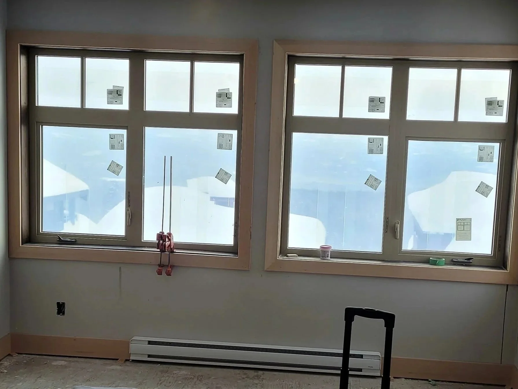 Interior room with two large double-pane windows with stickers, and a baseboard heater below. Construction tools and materials are seen, including a clamp, small cup, and green tape on the windowsill. The floor is under construction and unfinished.