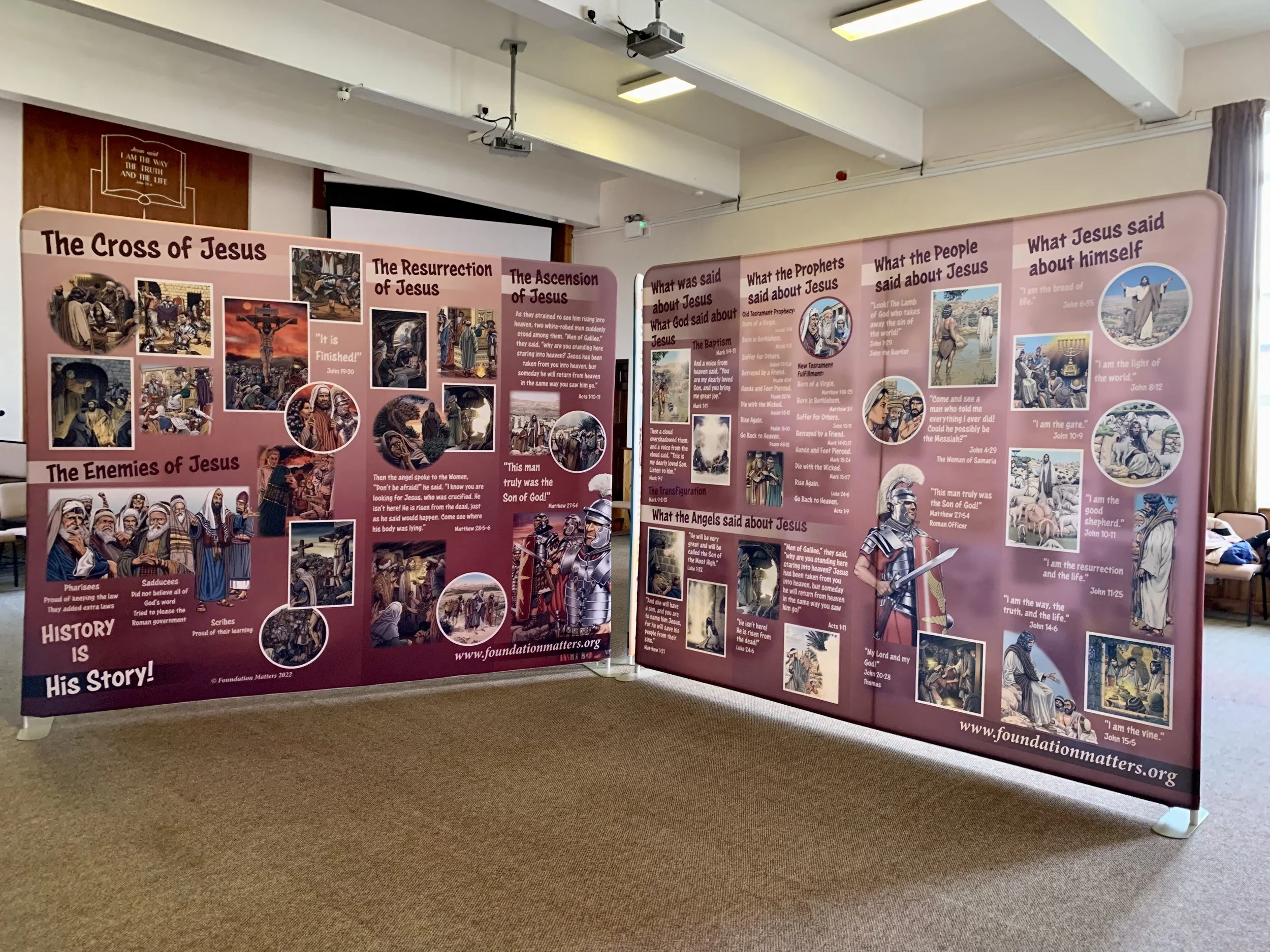 An indoor display with educational posters about the life of Jesus Christ, including images of biblical scenes, with titles such as 'The Cross of Jesus,' 'The Resurrection of Jesus,' and 'The Ascension of Jesus.' The posters contain text, illustrations, and quotes from the Bible, set in a room with a carpeted floor, white walls, and ceiling lights.