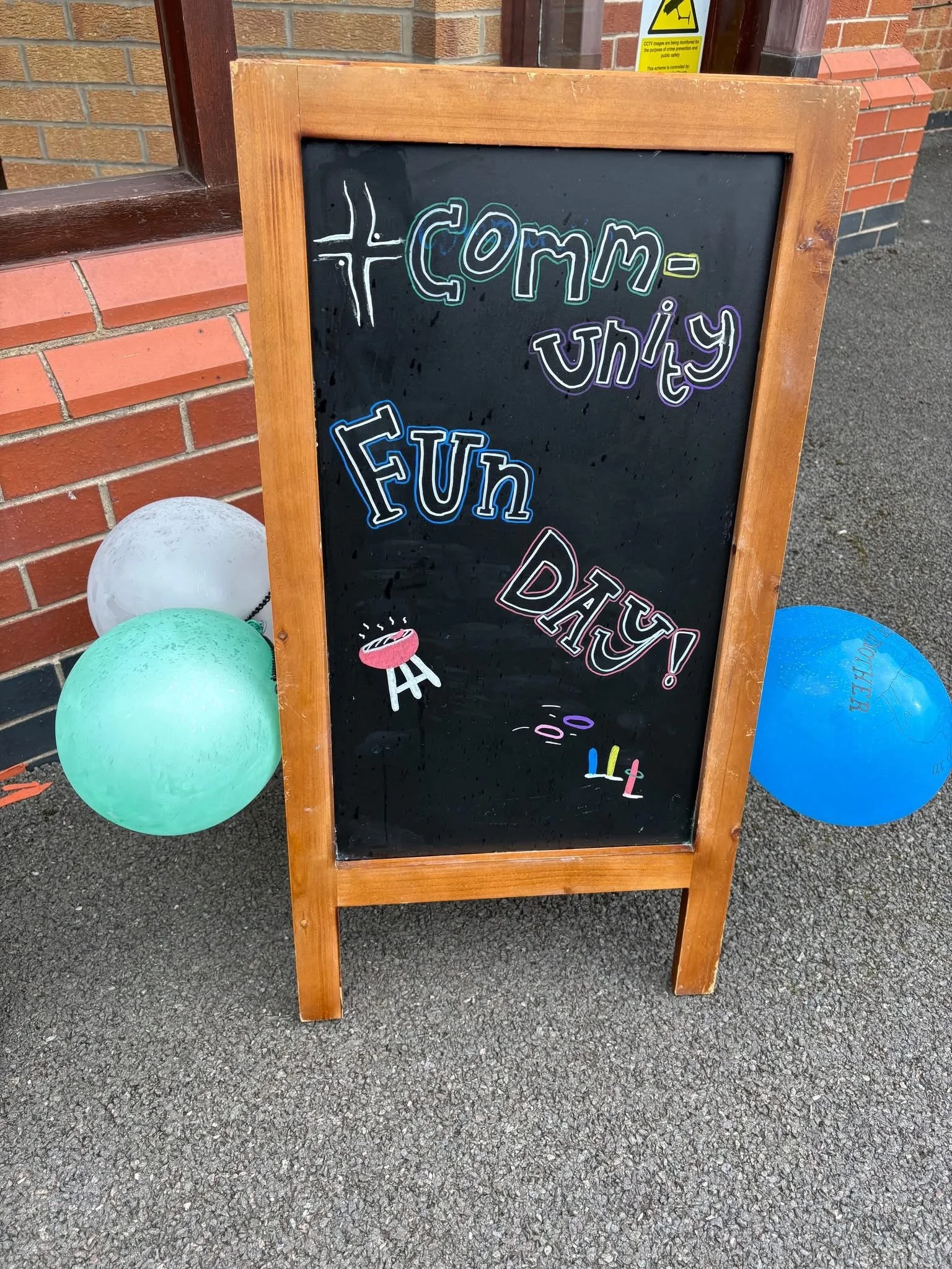 A black chalkboard sign with colorful writing that says 'Come enjoy' and 'Day!', decorated with balloons at the bottom, standing outside near a brick wall and sidewalk.