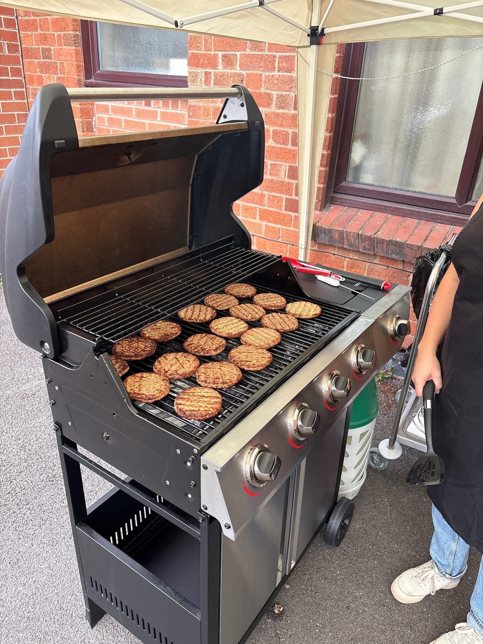 A person in jeans and white sneakers standing beside a barbeque grill with burgers cooking on it, located outdoors near brick walls and windows, under a canopy.