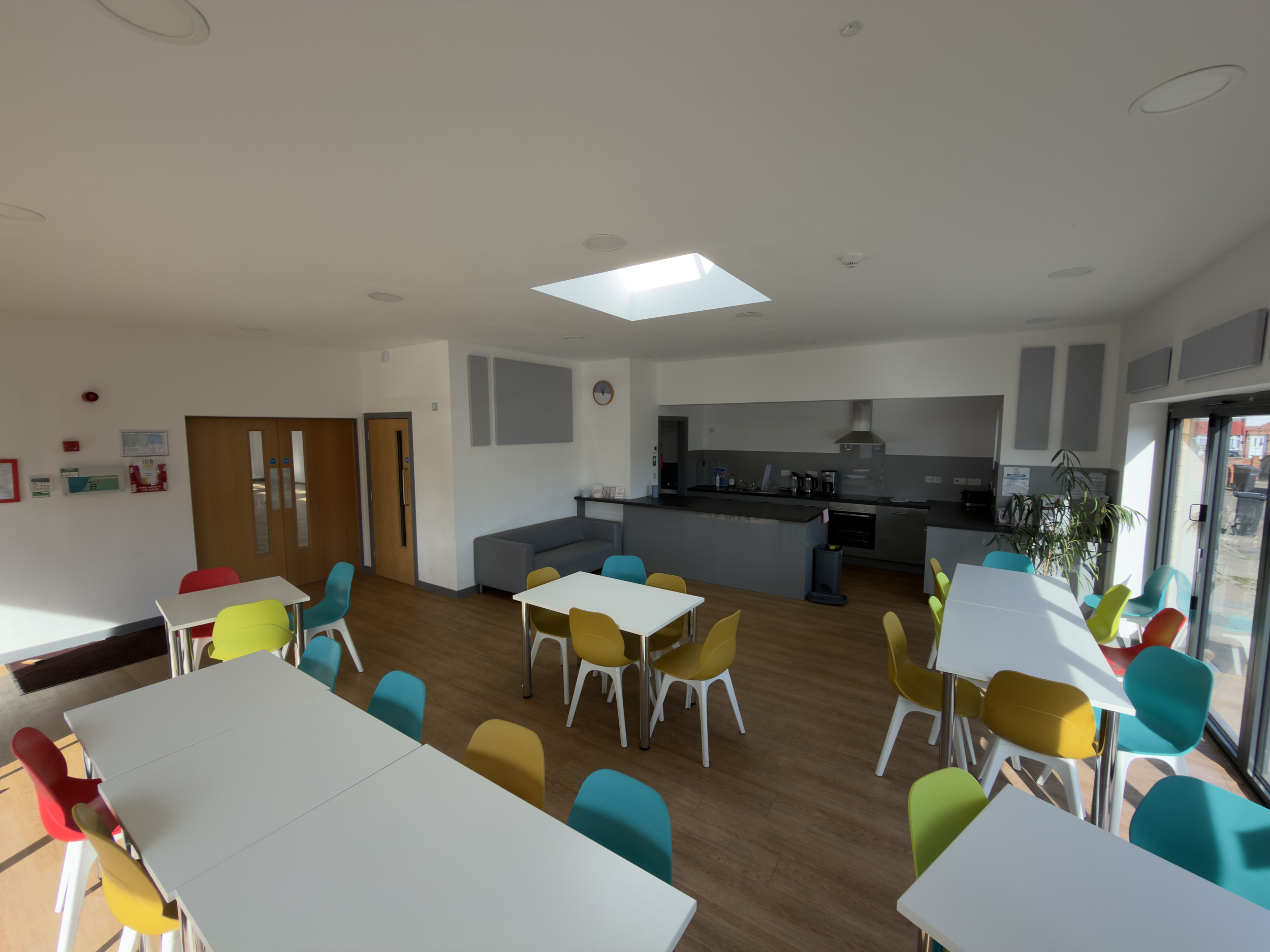 A spacious, modern room with white walls and ceiling, featuring a skylight letting in sunlight. The room has multiple white tables surrounded by colorful chairs in red, yellow, and teal. In the background, there's a kitchenette with gray cabinets, a stove, and a small gray sofa near the wall. A large window and potted plants are also visible.