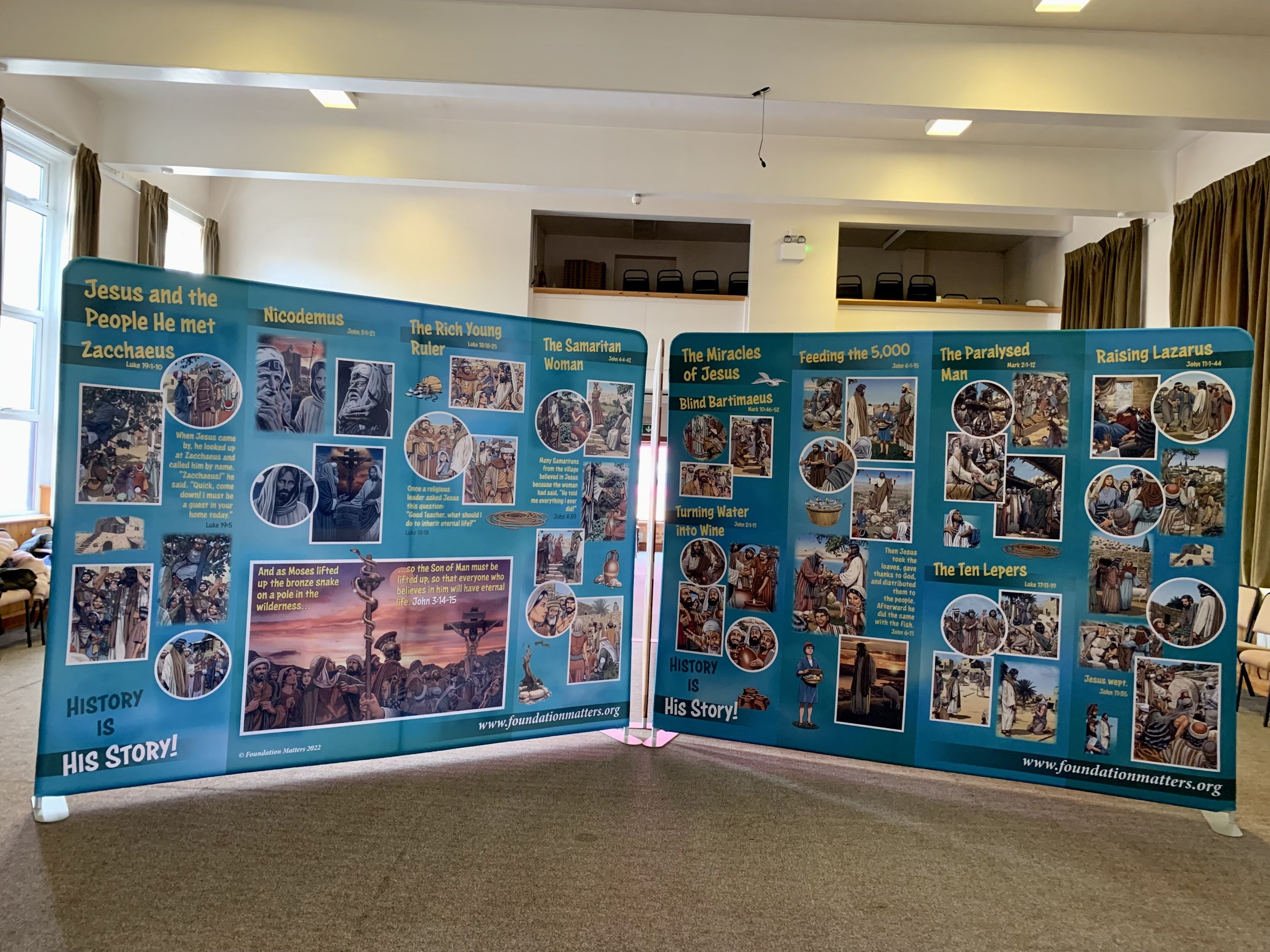 Exhibit panels with biblical artwork and text in a room with windows and curtains, discussing the story of Jesus and his followers.