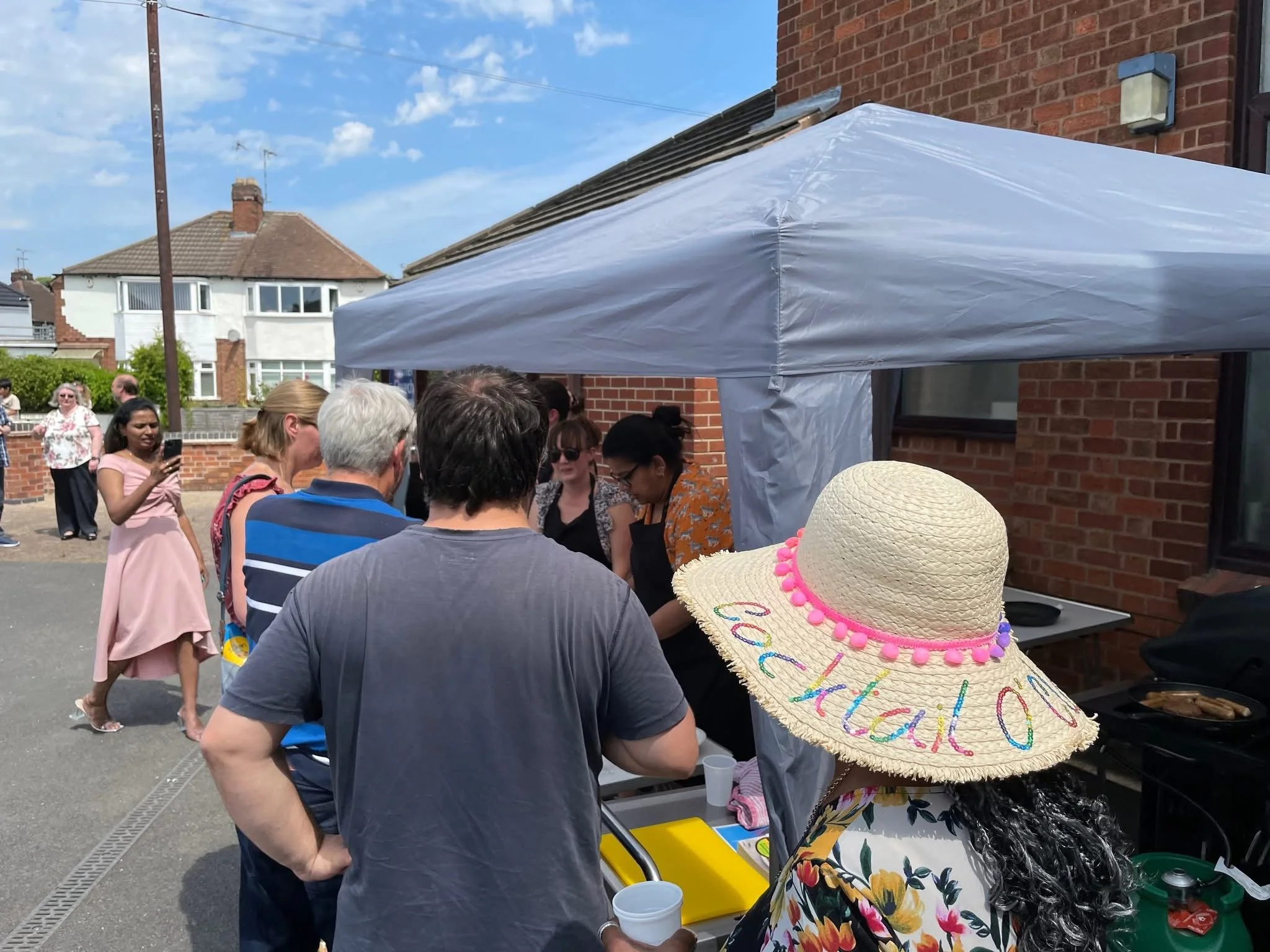 People gathered outdoors under a white canopy, with a woman wearing a large straw hat embroidered with colorful words. The background shows a brick building and other residential houses under a blue sky with scattered clouds.