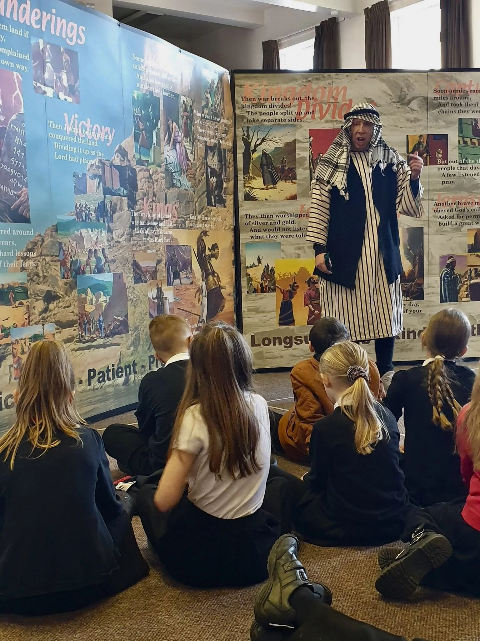A classroom scene with children sitting on the floor, attentively listening to a person dressed in biblical costume standing in front of large illustrated story posters about ancient biblical events.
