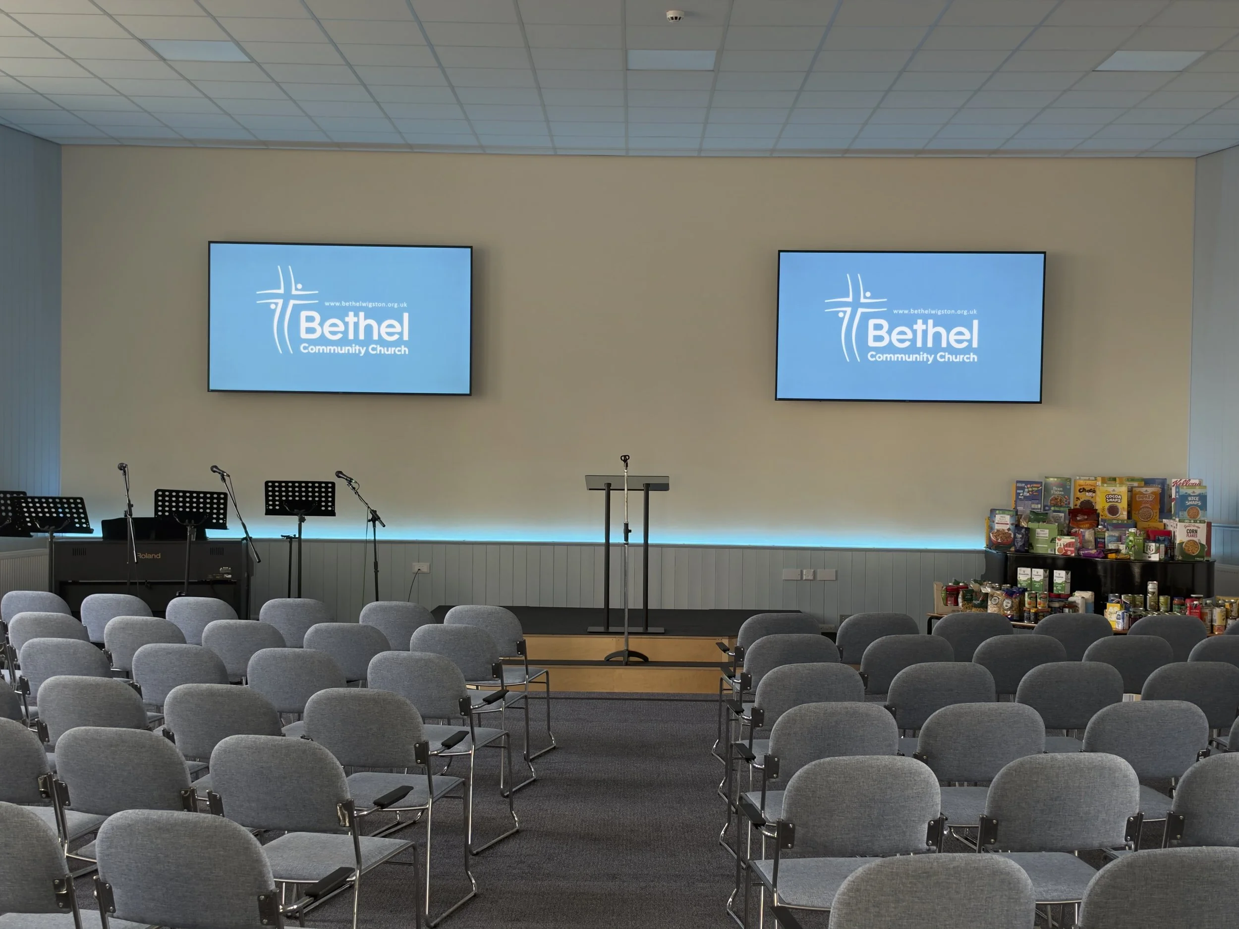 Bethel Community Church sanctuary with rows of gray chairs, pulpit, microphones, music stands, dual projection screens, and food donation display