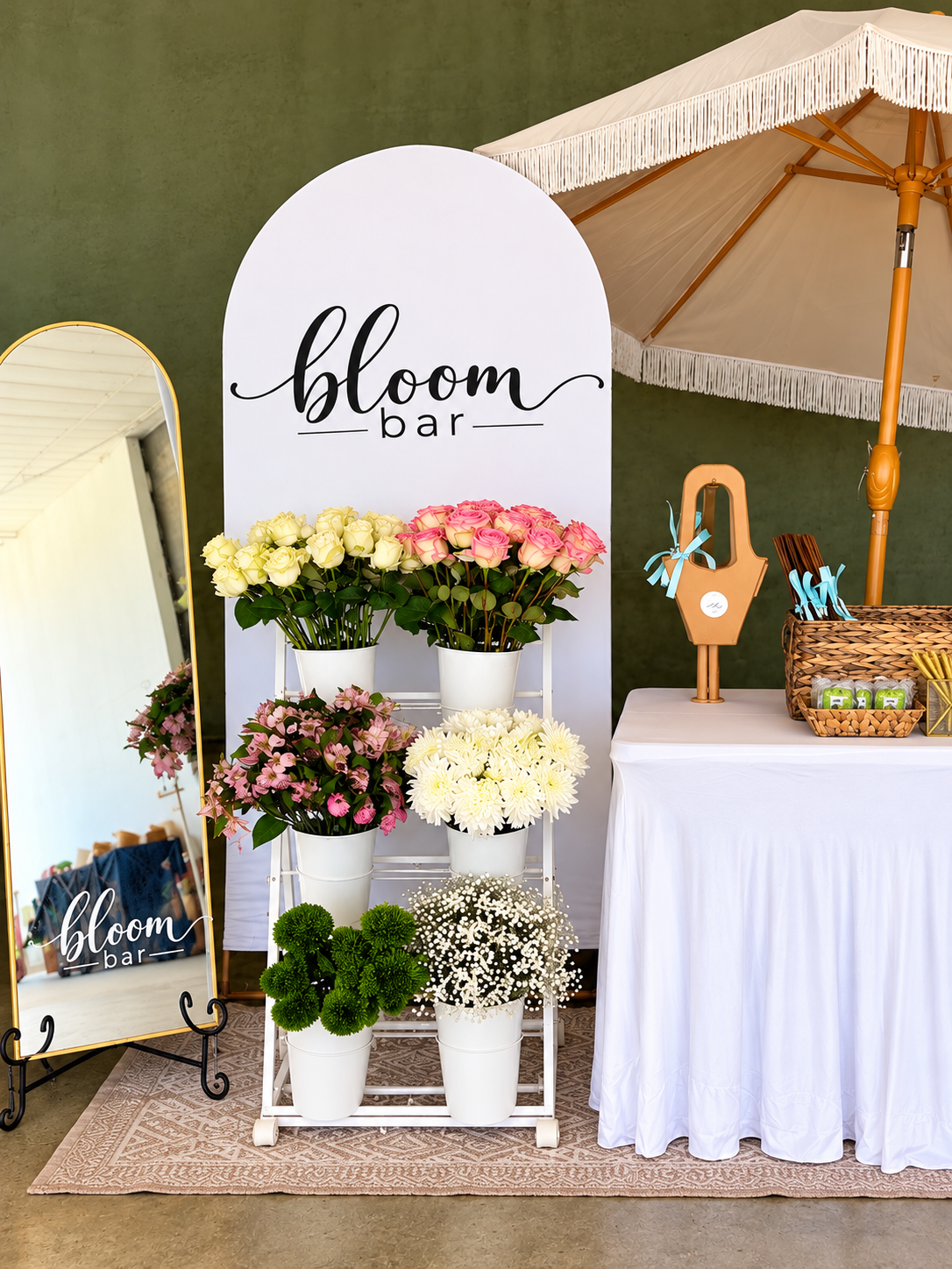 BloomMuseCo Bloom Bar display with pink and ivory flowers, a white arched sign, gold mirror and cream fringe umbrella