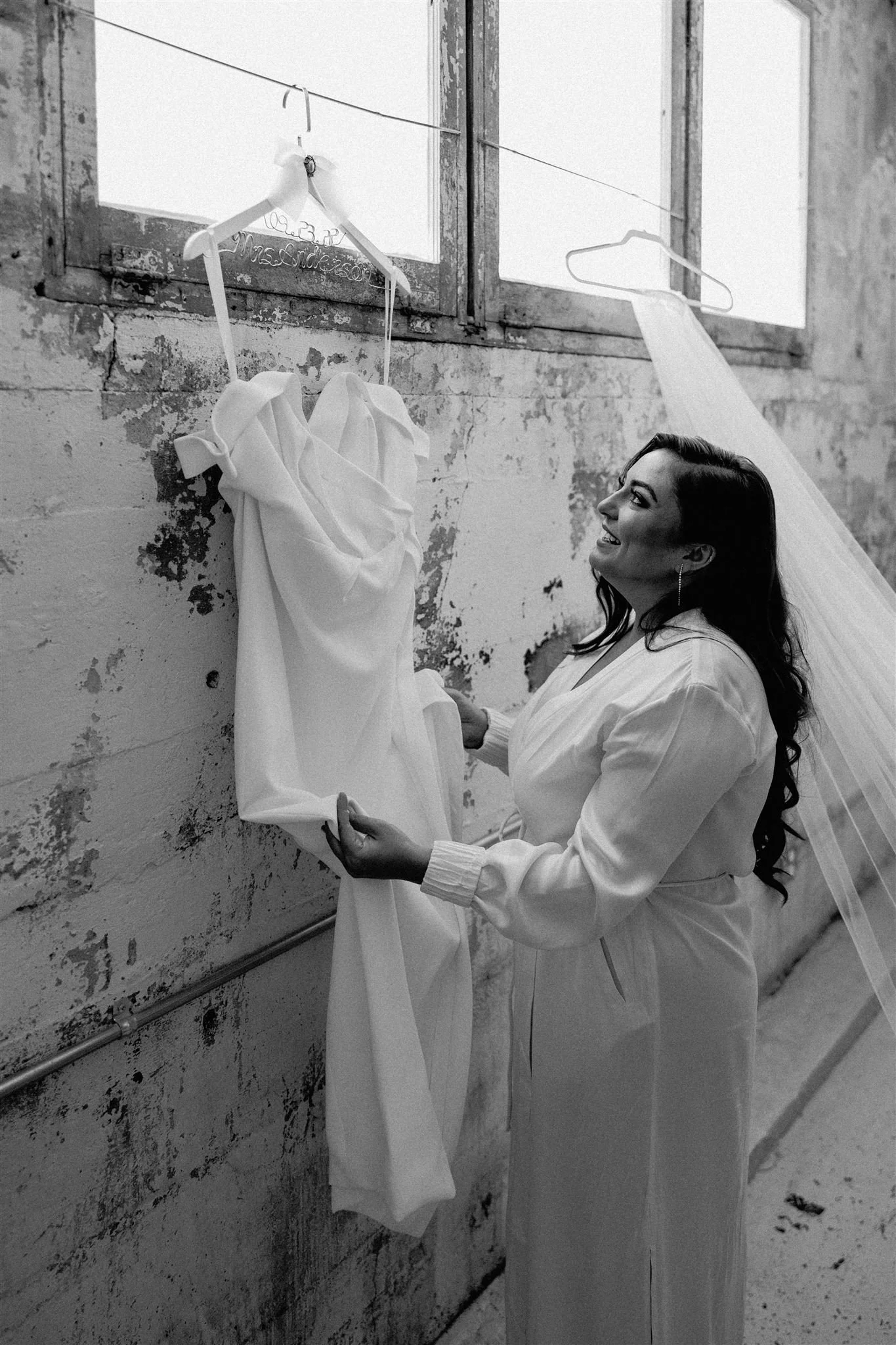 A woman in a white shirt holding a wedding dress, smiling as she looks at it, with the dress hanging on a hanger. The hanger has the name 'Madison' written on it. The setting appears to be an industrial space or studio with a distressed wall and a wi