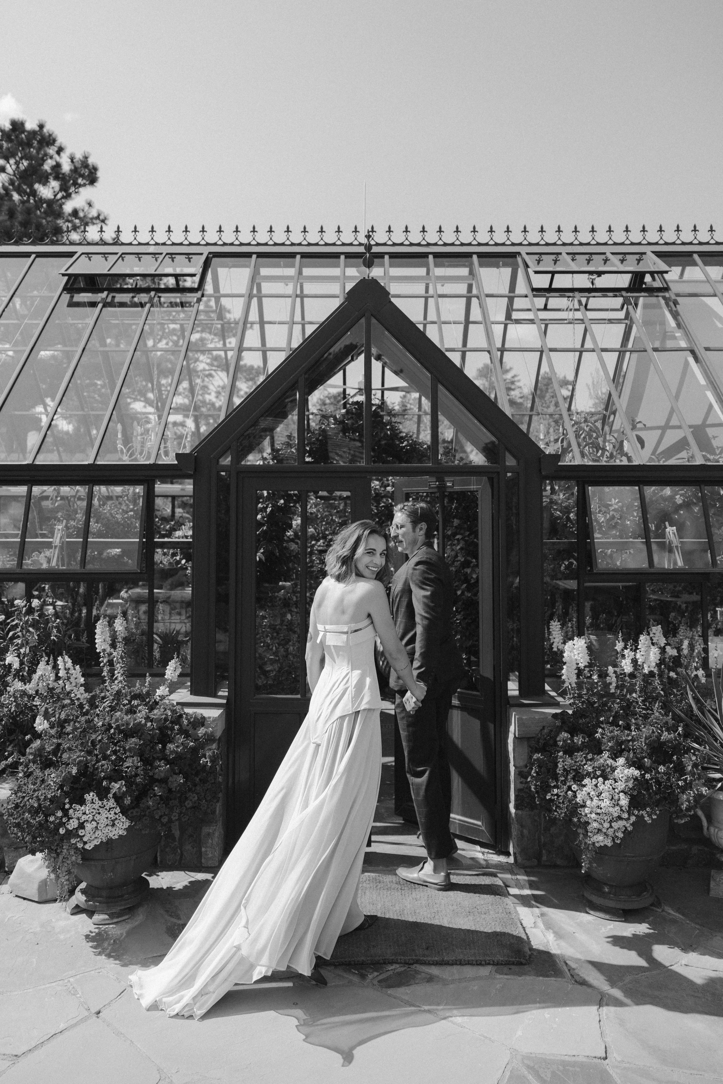 A smiling bride and groom holding hands and looking at each other in front of a glass greenhouse building with flowers in planters on either side.