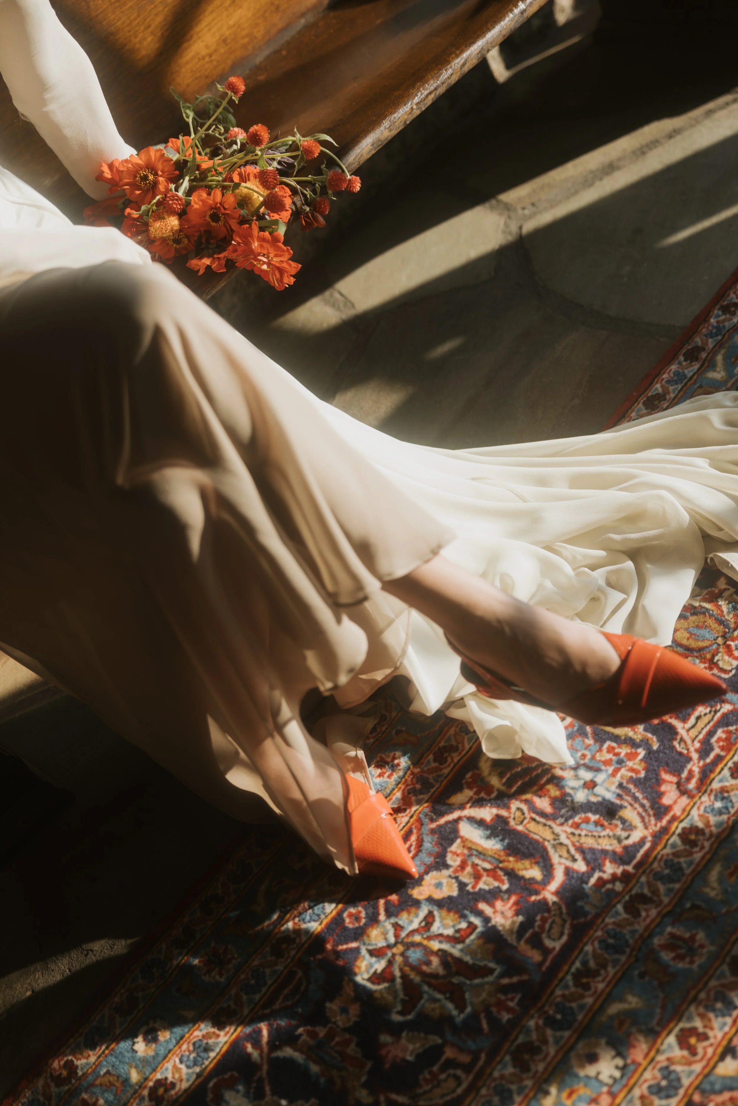 The image shows a person sitting on a Persian rug, wearing beige shoes, a white dress or skirt, and orange socks. They are holding a bouquet of orange flowers with some round orange seed pods. The person is near a wooden bench, and the lighting suggests natural sunlight.