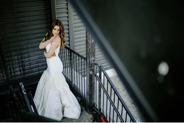 A woman in a strapless white dress with a ruffled skirt on a fire escape, looking down with her arms crossed over her chest.