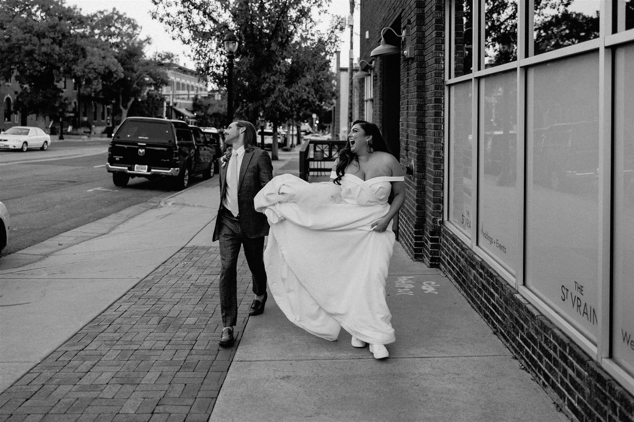 A bride in a wedding gown walking on a city sidewalk with a man in a suit, both laughing. The bride is holding part of her dress, and there are parked cars, trees, and buildings in the background.