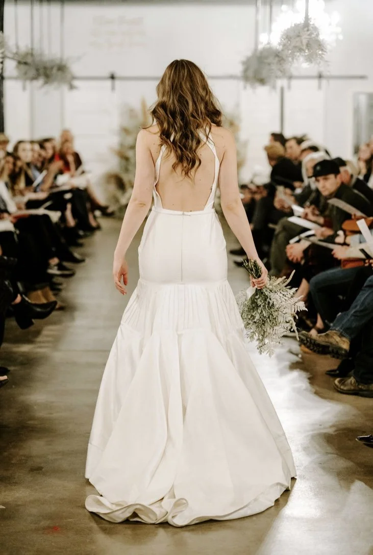 A woman in a white wedding dress walking down a runway holding a bouquet of flowers during a fashion show surrounded by seated audience members.