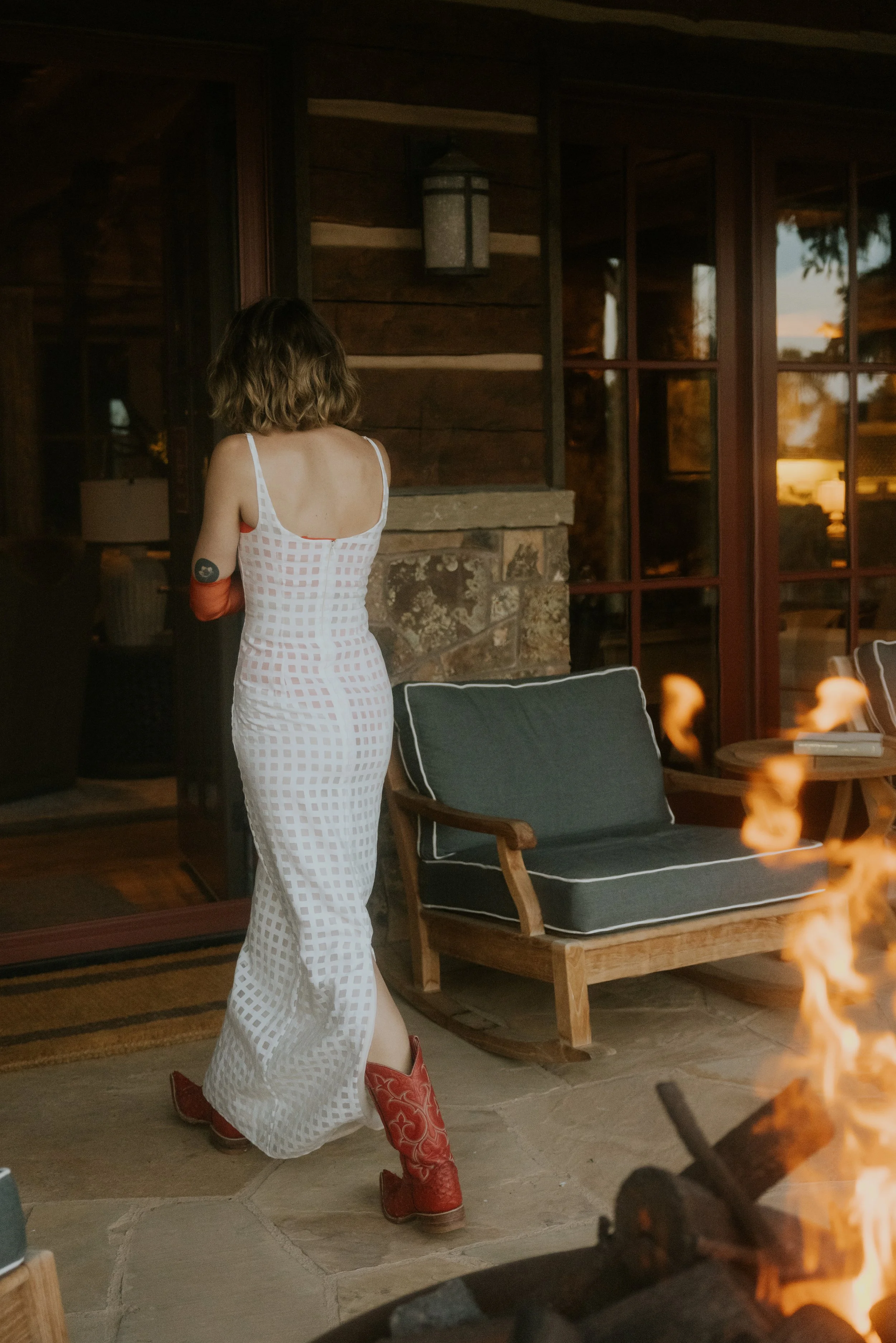 A woman in a white checkered dress with red cowboy boots standing near a fireplace in a rustic wooden room with large windows at sunset.
