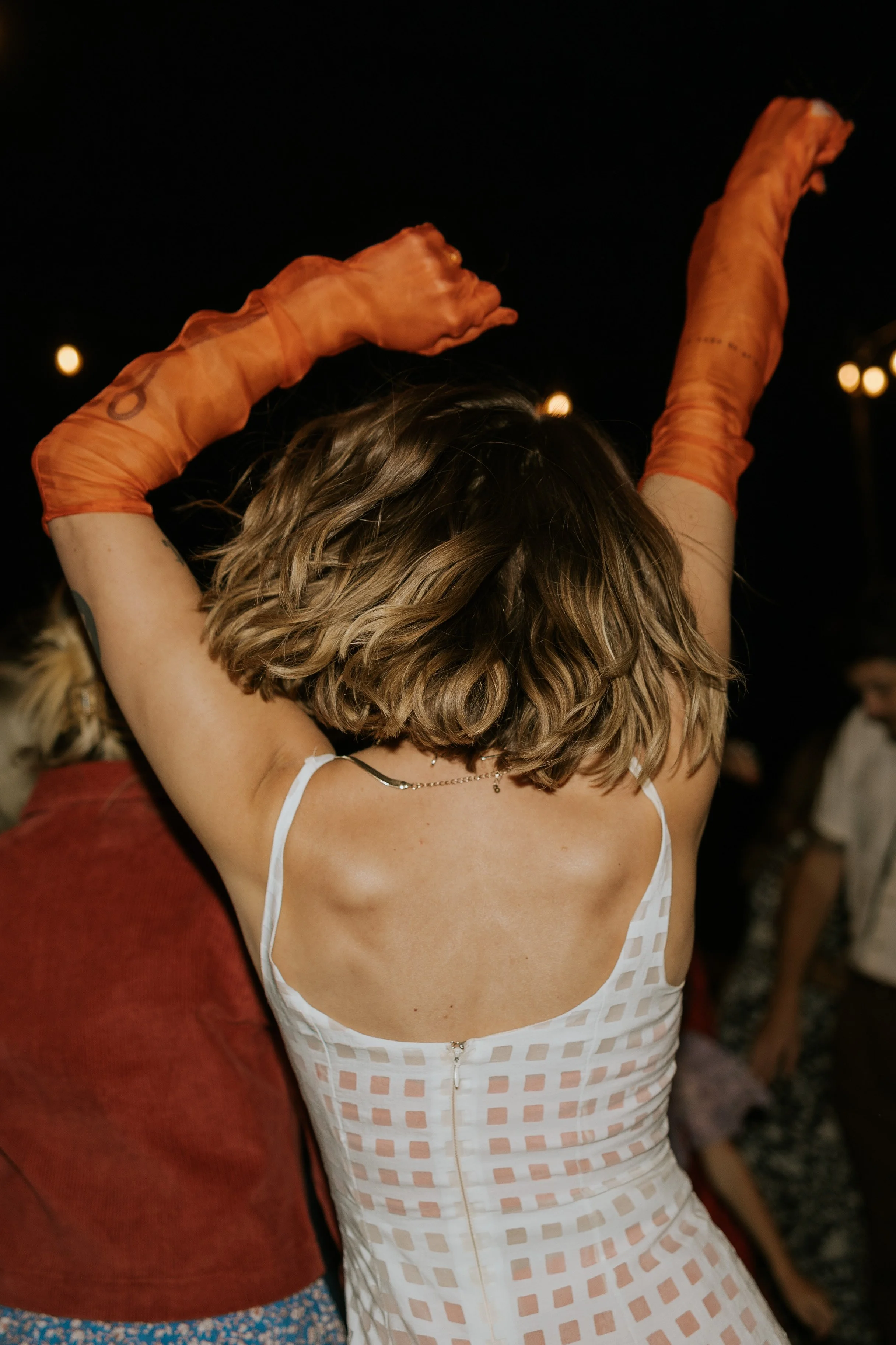 A woman in a white checkered dress with spaghetti straps dancing at night, wearing orange gloves, with her body facing away from the camera.