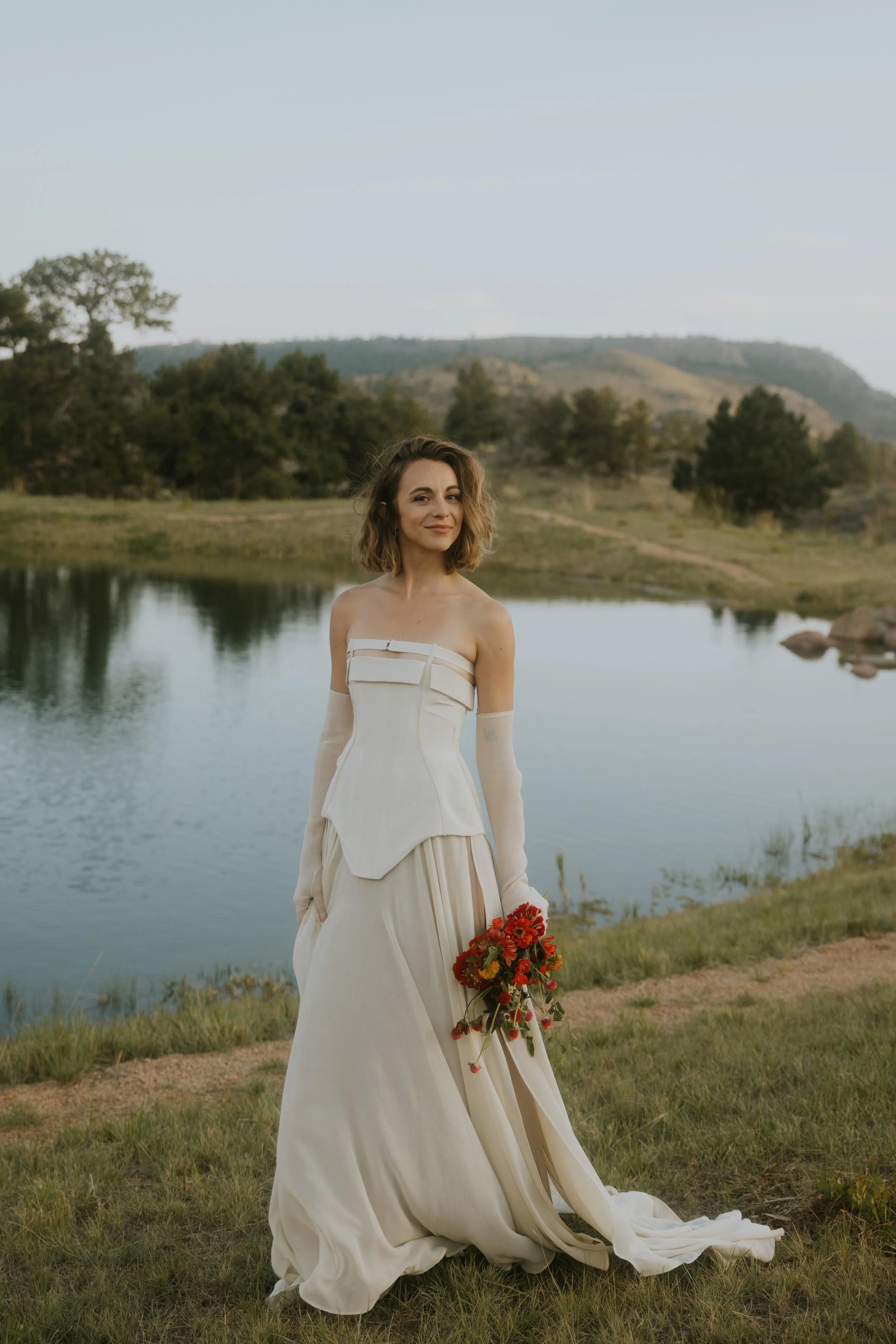 A woman in a white wedding dress standing outdoors near a pond, holding a bouquet of red flowers, with hills and trees in the background.