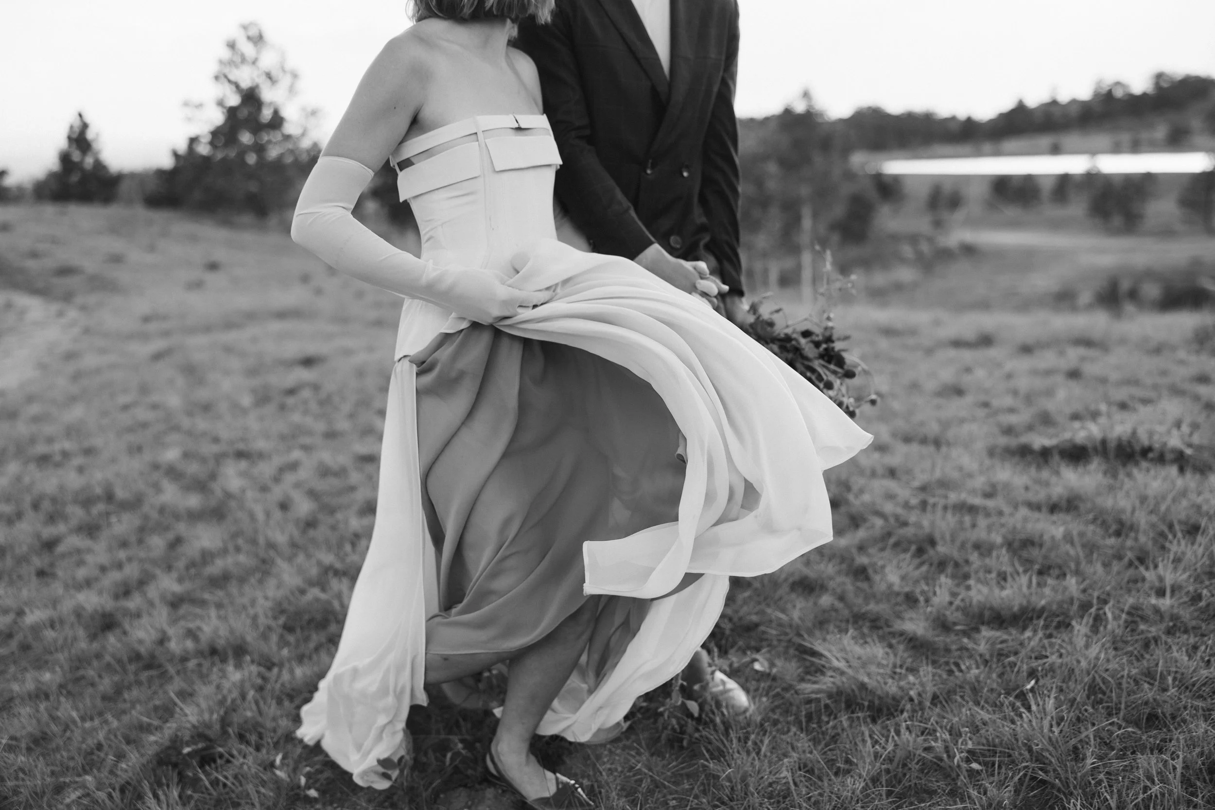 A black and white photo of a bride in a strapless wedding gown holding up her dress in an outdoor grassy field, with a man in a dark suit standing beside her. The bride's face is not visible.