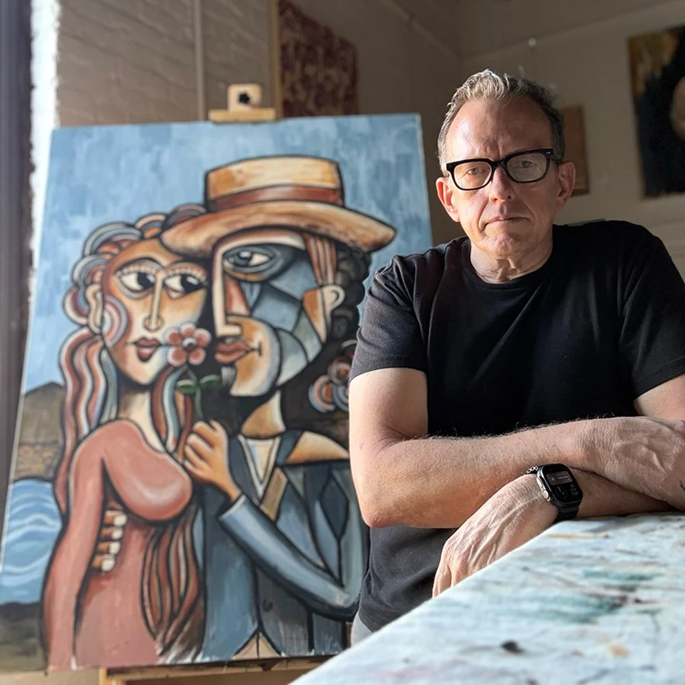A man with glasses and a black t-shirt sitting at a table next to an abstract painting of two people, one wearing a hat, painted in a Cubist style.